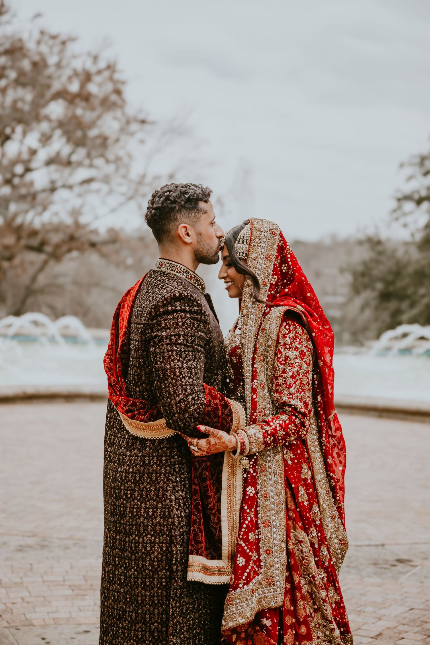 A couple in traditional South Asian wedding attire share an intimate moment, with the bride wearing a red embroidered lehenga.