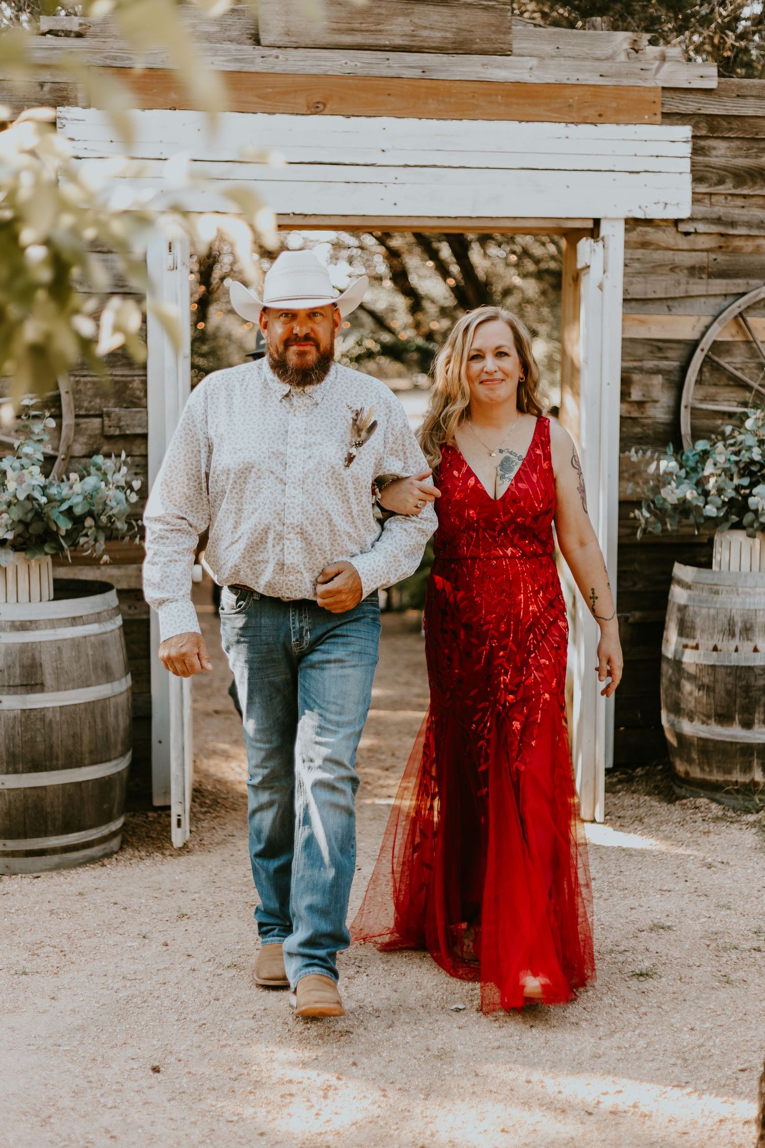A couple walks together at a rustic venue with wooden barrels and an arbor, the woman wearing a sparkly red gown.
