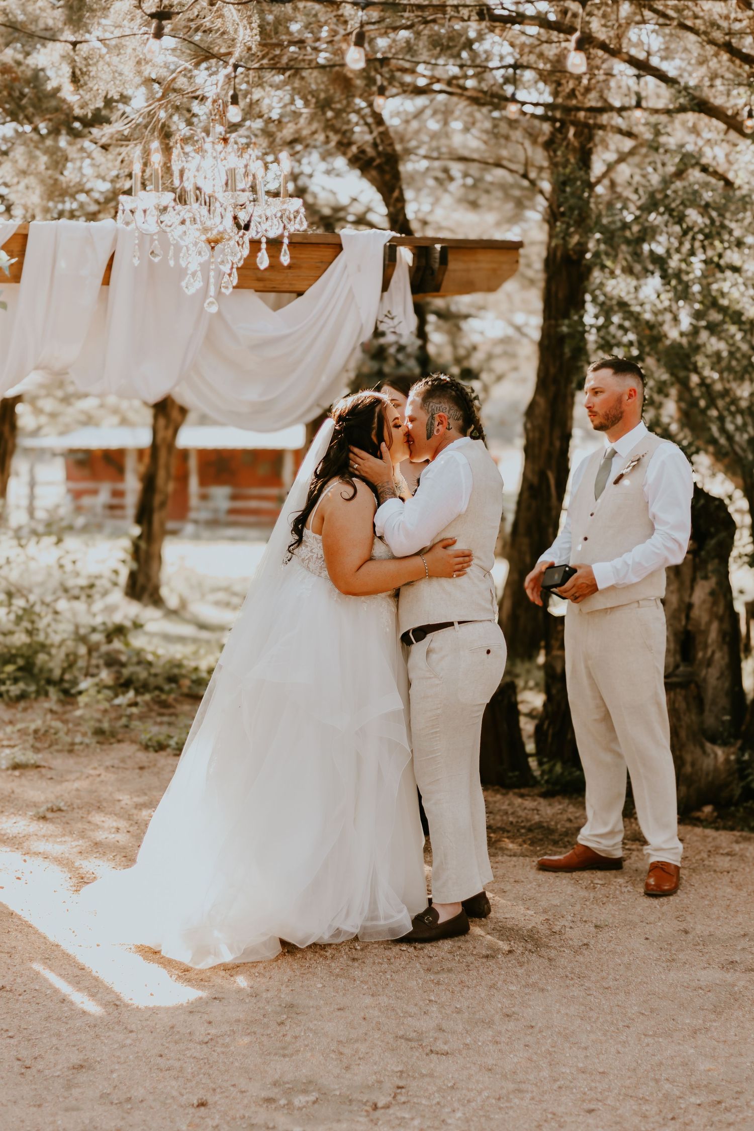 Rustic outdoor wedding ceremony with white decor, trees, and a wooden cabin in the warm sunlit background.