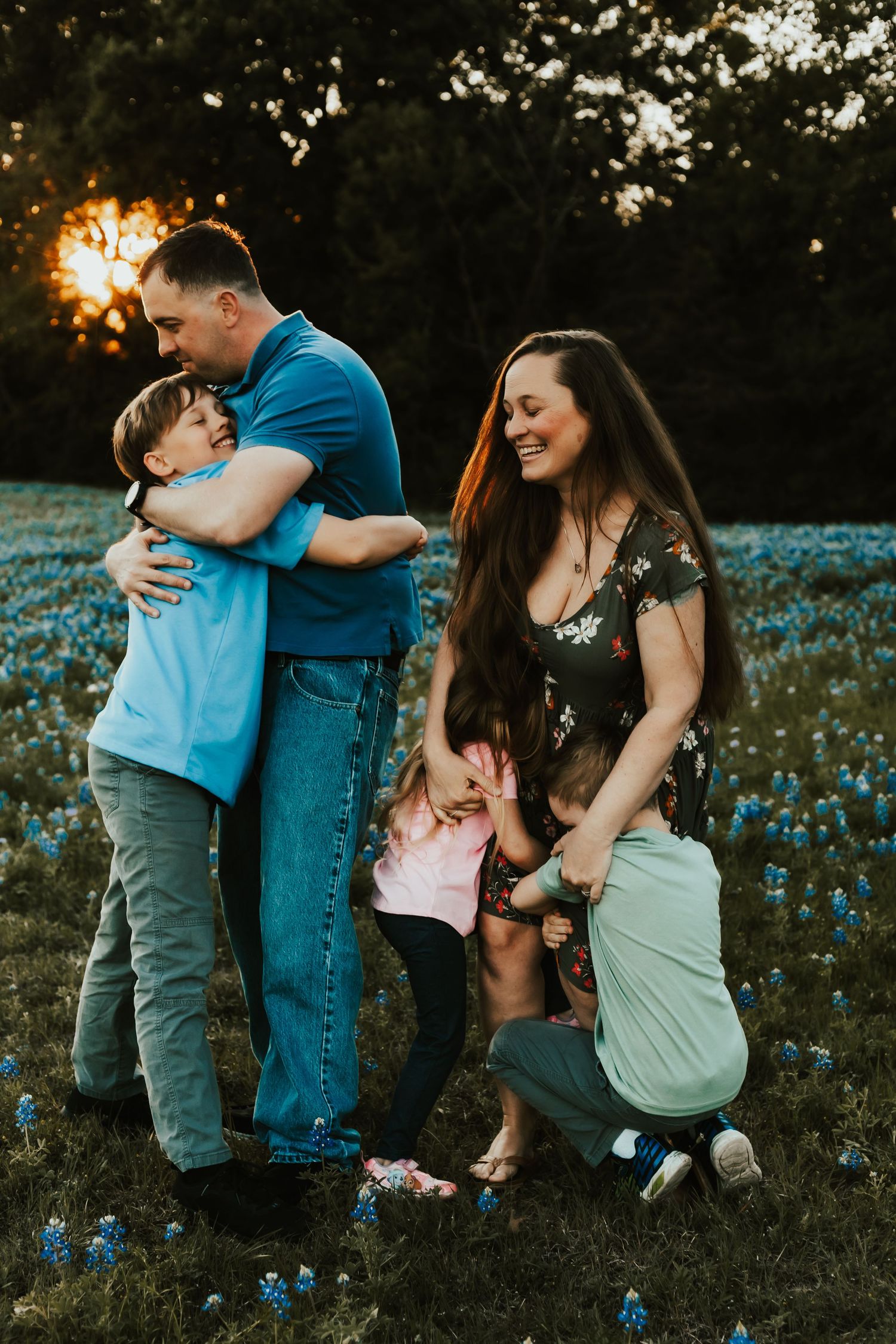 A family stands together in a field of bluebonnets at sunset with warm golden light filtering through the trees.