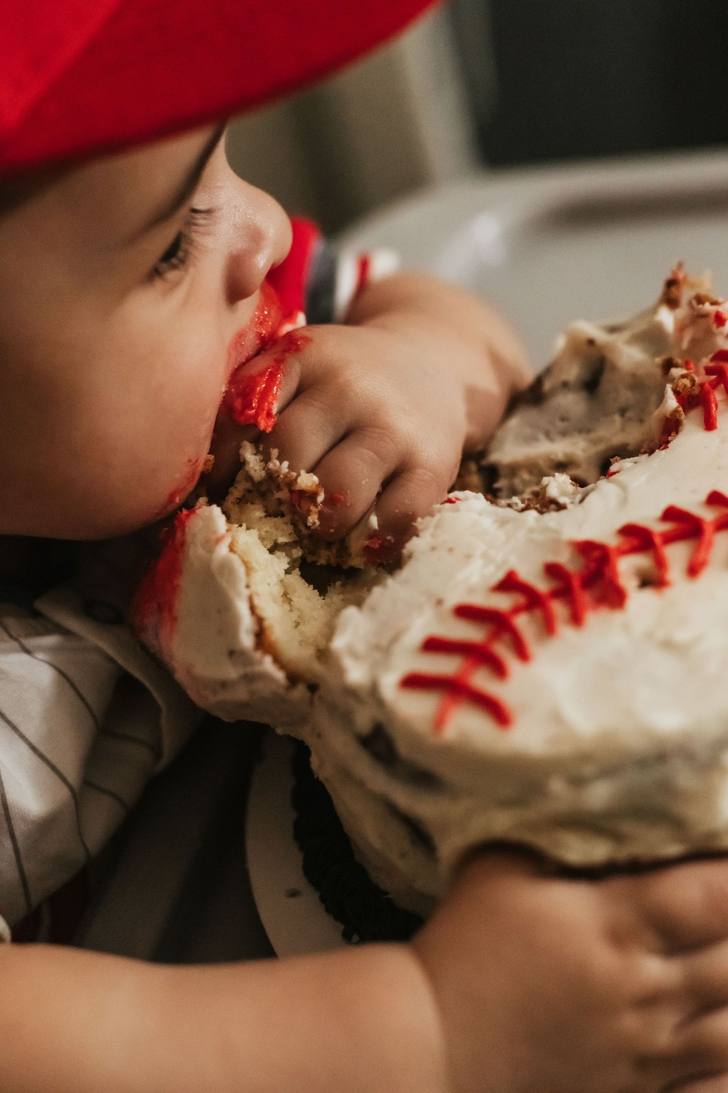A young child with frosting on their face eagerly devours a baseball-themed cake with red stitching decorations.