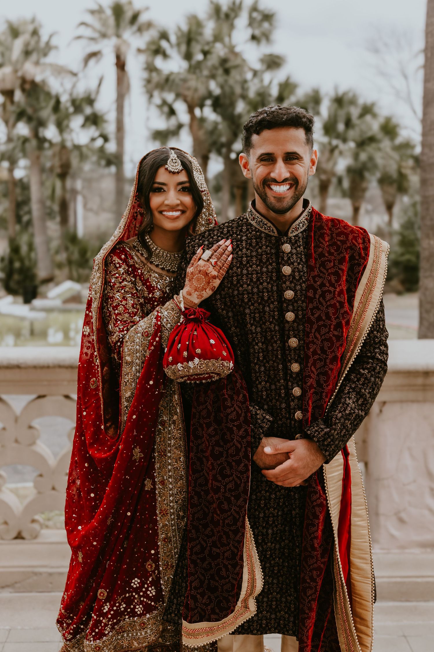 Two people in traditional Indian wedding attire with red and gold embroidered outfits pose together outdoors near palm trees.