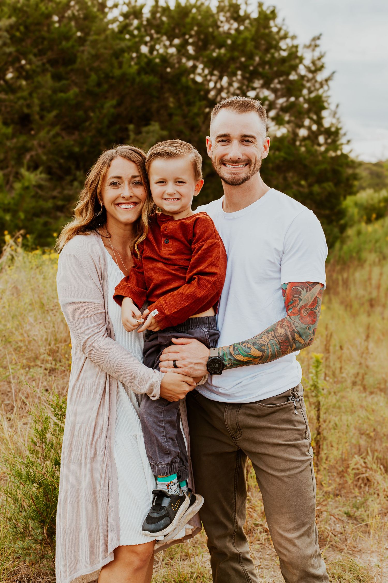 A family poses together outdoors in a grassy field at golden hour with trees in the background.