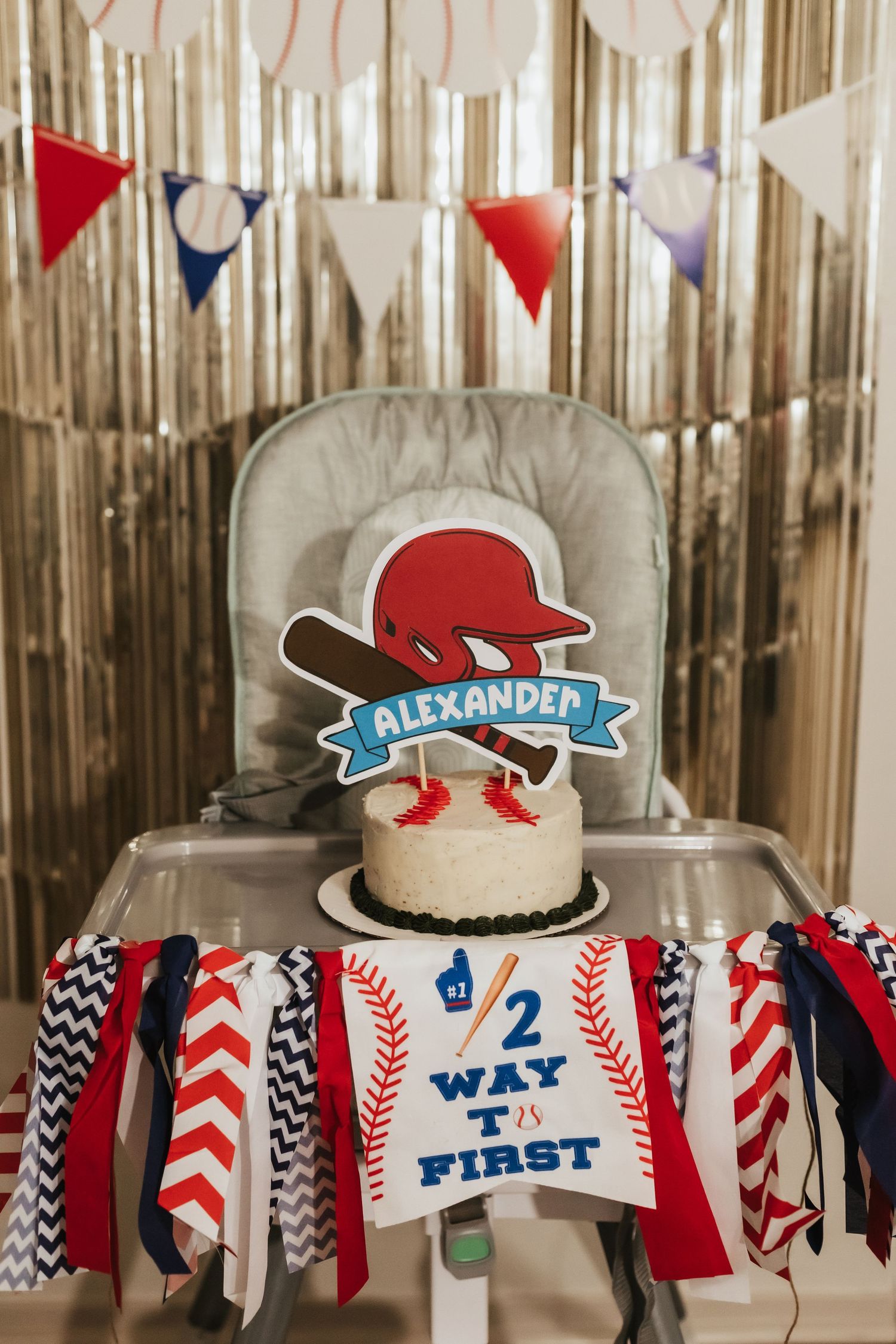 A decorated high chair with red and navy bunting, baseball themed decor, and a 'First' birthday sign for Alexander's celebration.