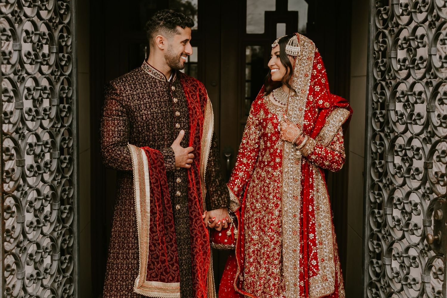 Traditional South Asian wedding couple wearing ornate red and brown embroidered wedding attire holding hands.