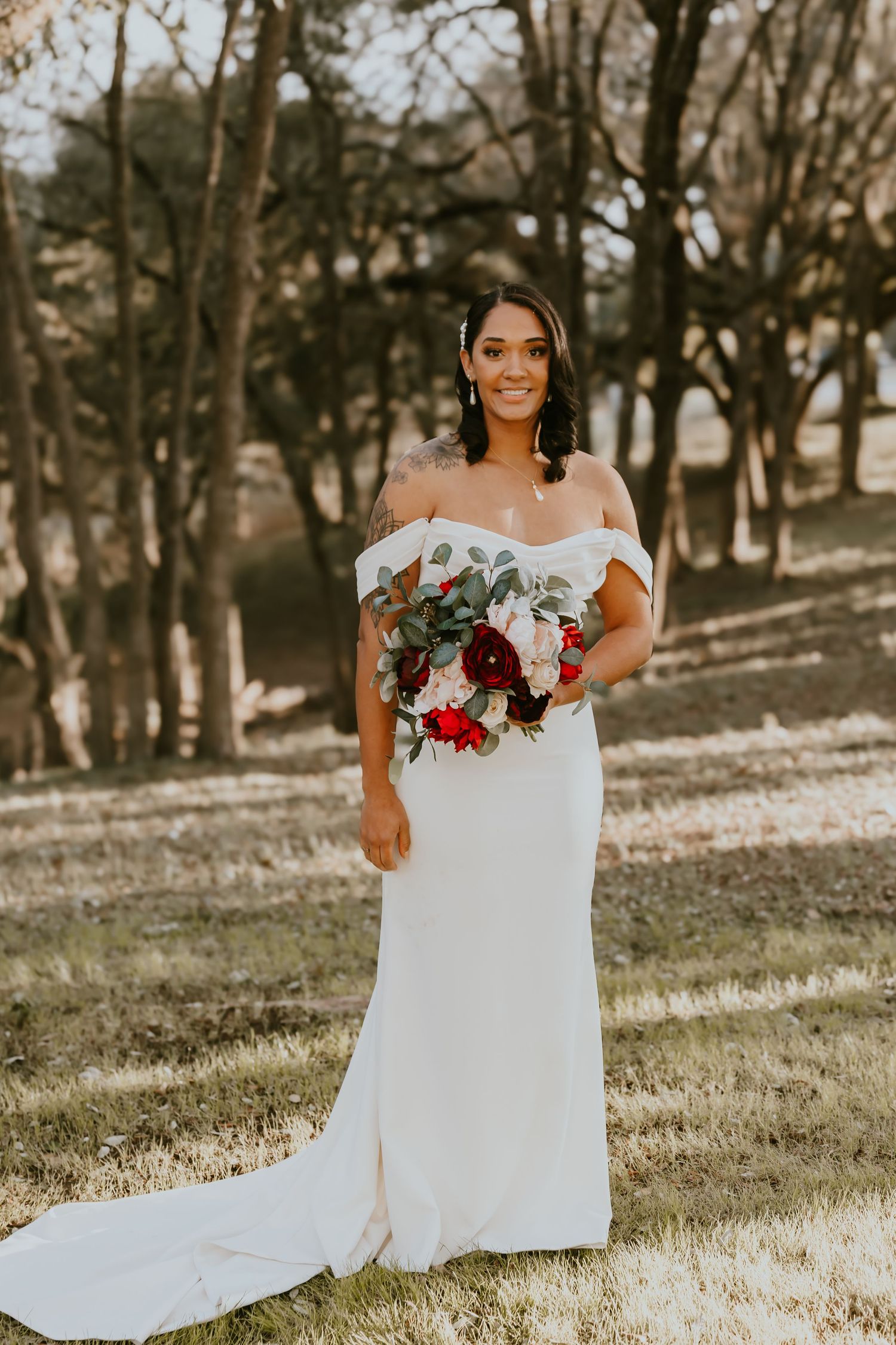 Elegant bride in off-shoulder white gown holds vibrant red and green bouquet while standing in sunlit outdoor setting.