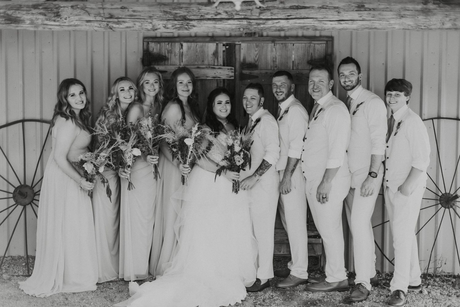 A black and white wedding party photo with bridesmaids in light gowns and groomsmen in light suits posed against a rustic wall.