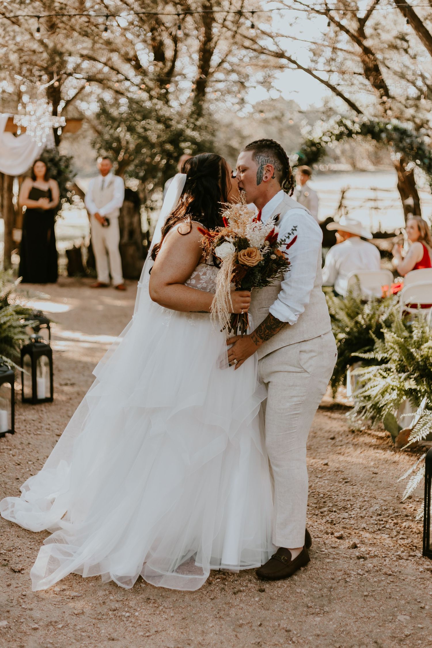A couple shares their first kiss at an outdoor wedding ceremony under trees with guests looking on in the background.