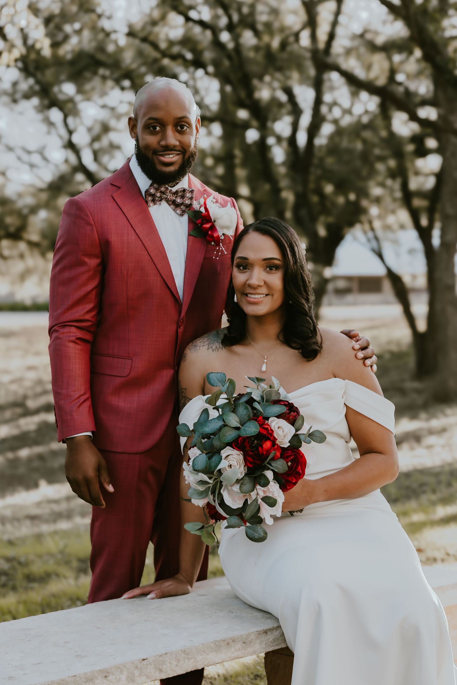 A beautiful outdoor wedding photo with the couple wearing a burgundy suit and white off-shoulder dress holding a floral bouquet.