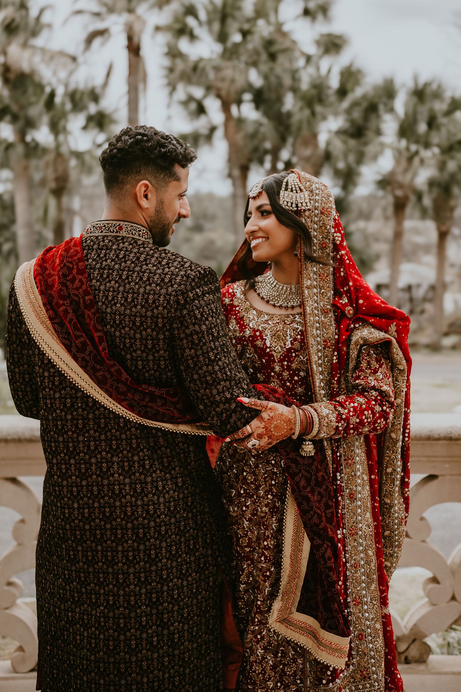 Traditional Indian wedding couple in ornate red and black outfits share an intimate moment against palm trees.