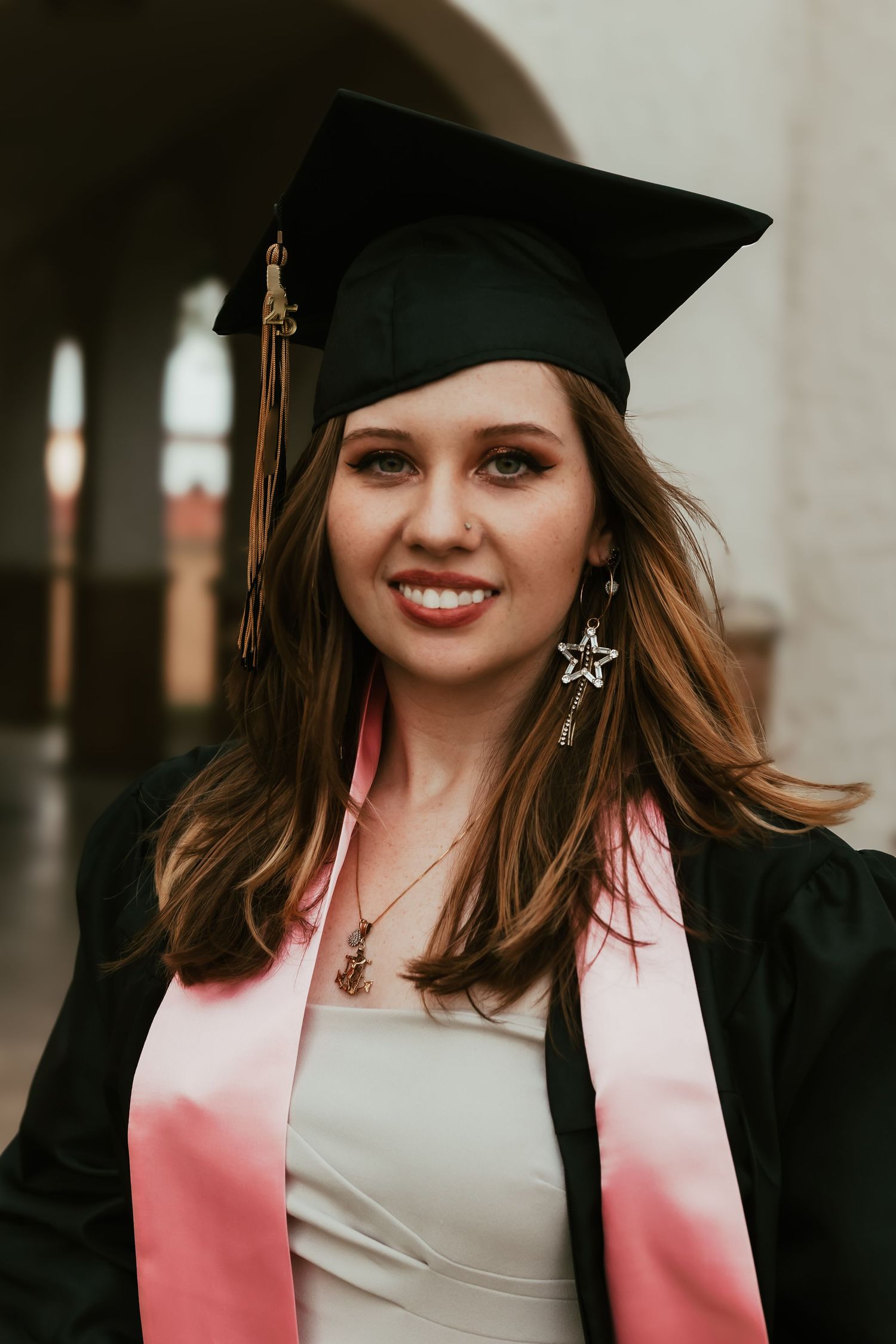 Graduate wearing black cap and pink stole smiles in graduation portrait photo series.