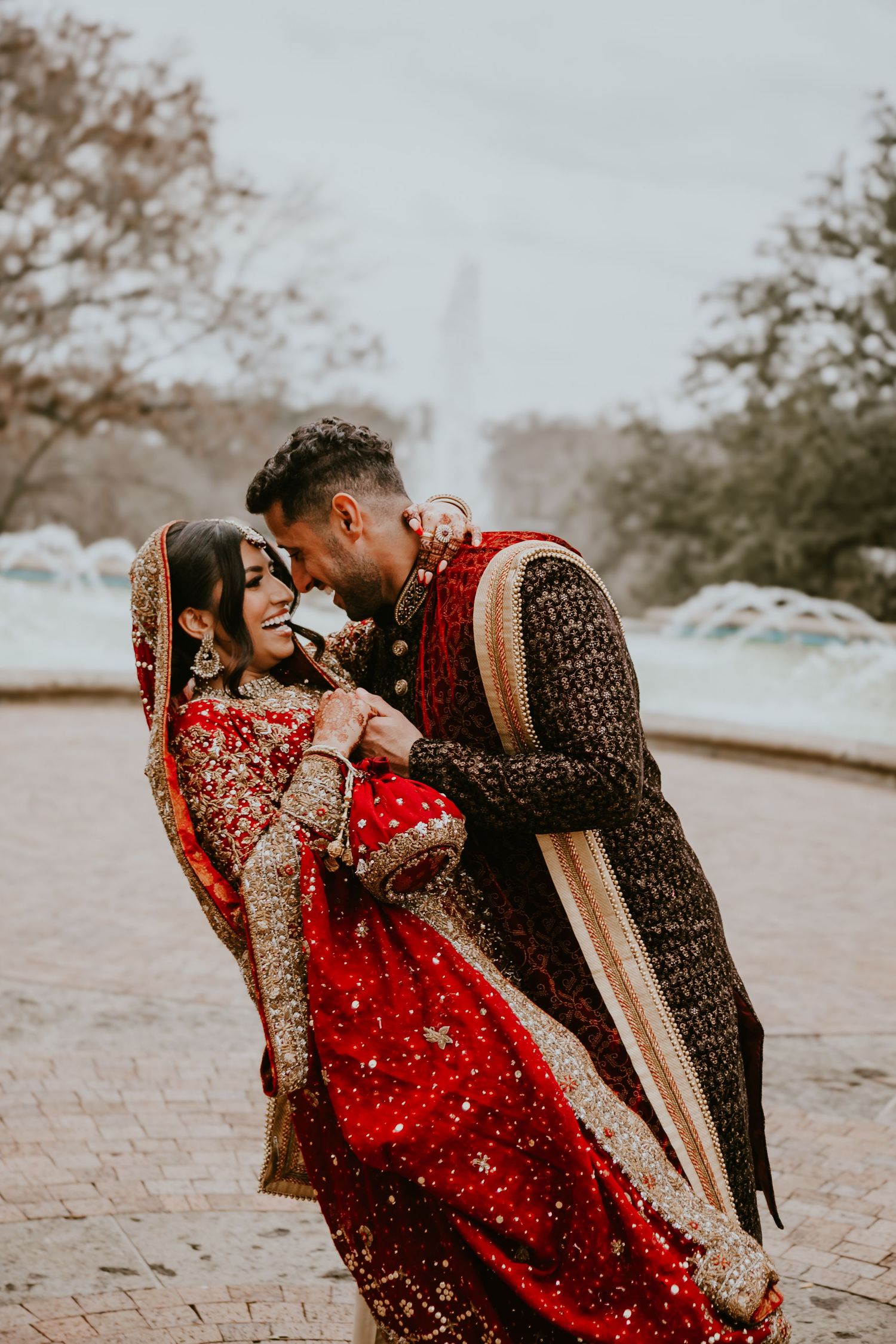 Couple in red and black traditional South Asian wedding attire share an intimate moment in a park during autumn.