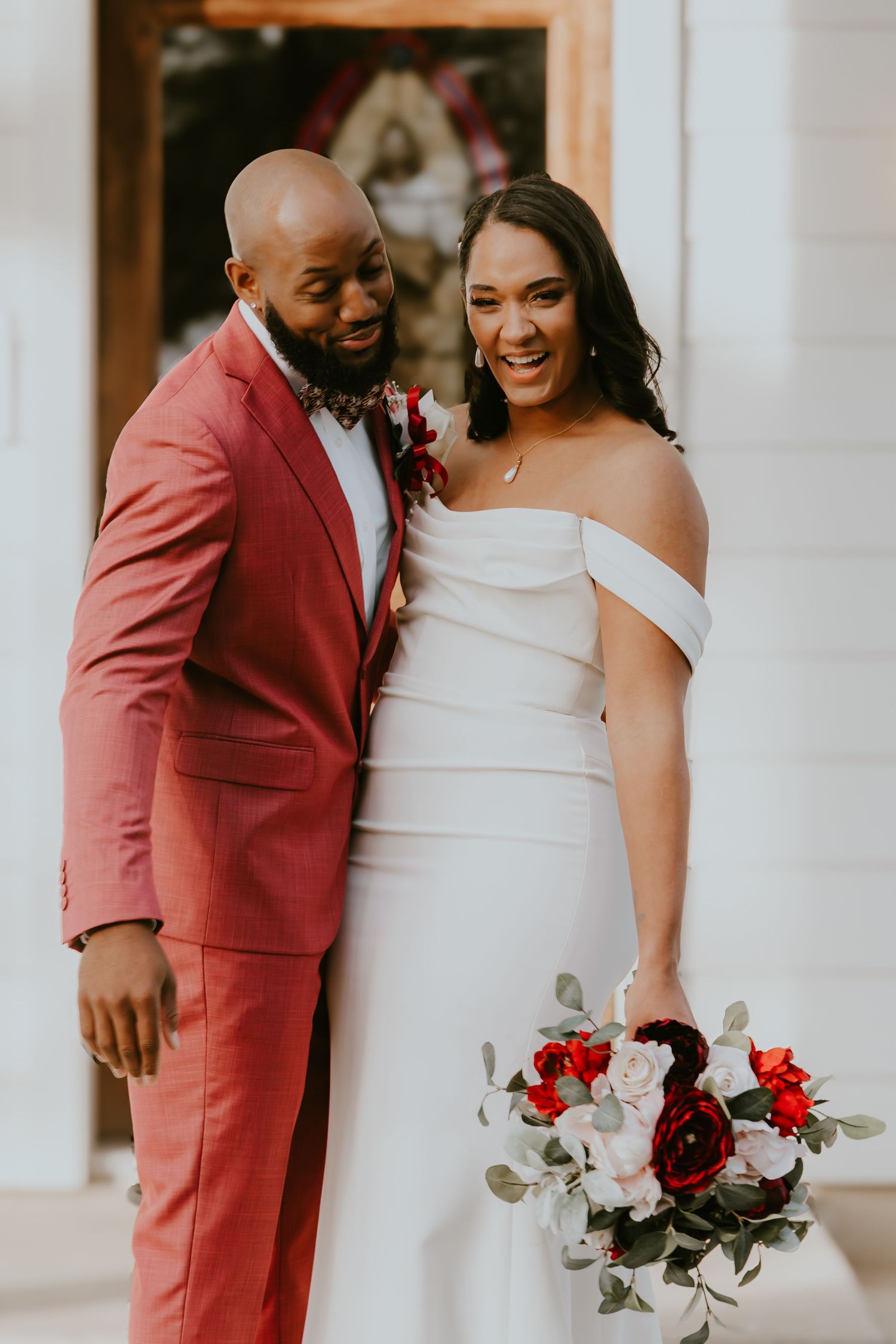 Happy newlyweds share a joyful moment in formal attire with a beautiful red and white floral bouquet.