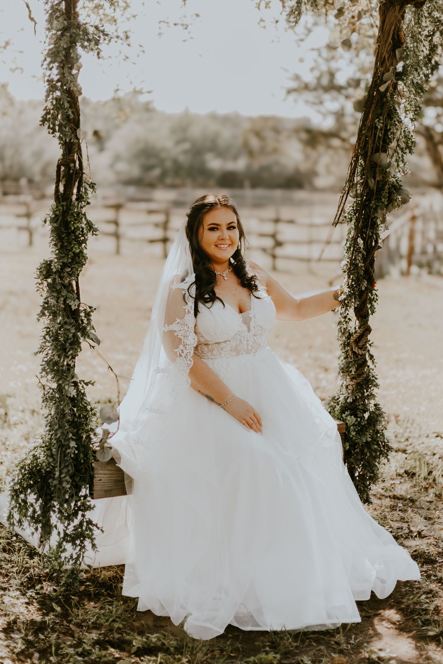A bride in a flowing white lace gown sits on a rustic swing adorned with greenery in a dreamy outdoor setting.