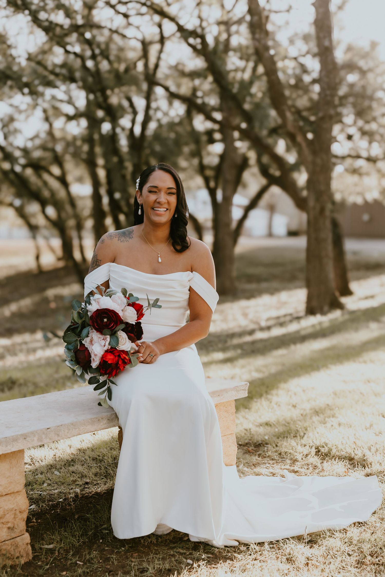 A bride in an elegant off-shoulder white gown holds a red and white bouquet while sitting outdoors during golden hour.