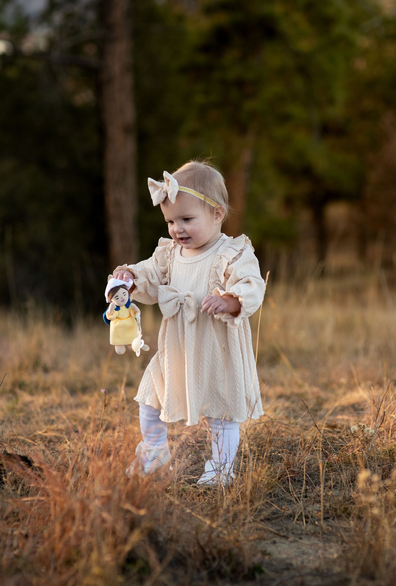 Toddler walking through grass carrying a doll wearing a bow