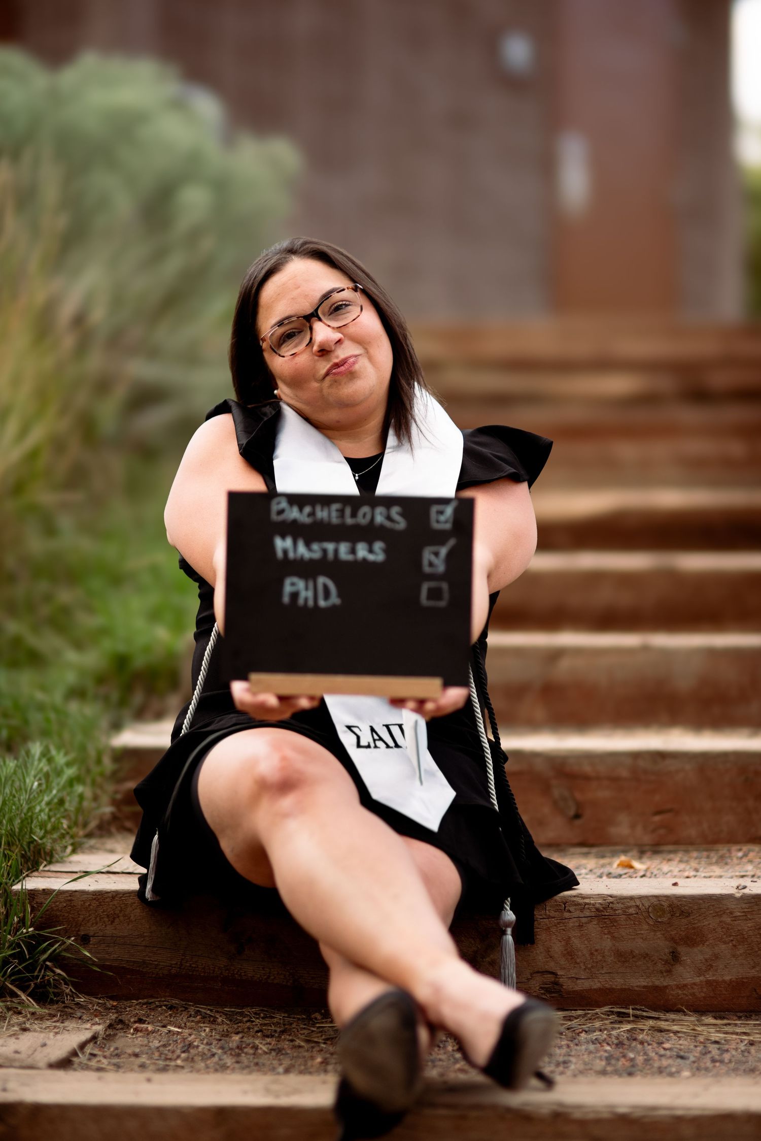 woman holding a chalkboard with checkmarks next to each completed degree while sitting on steps