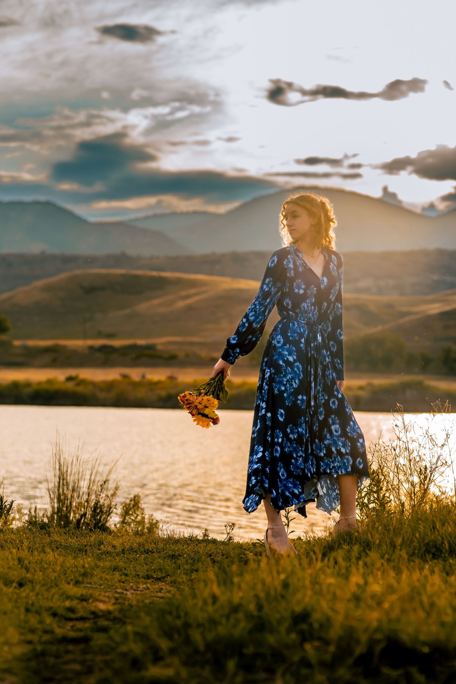 young woman standing in front of a lake holding flowers with mountains in the background at sunset
