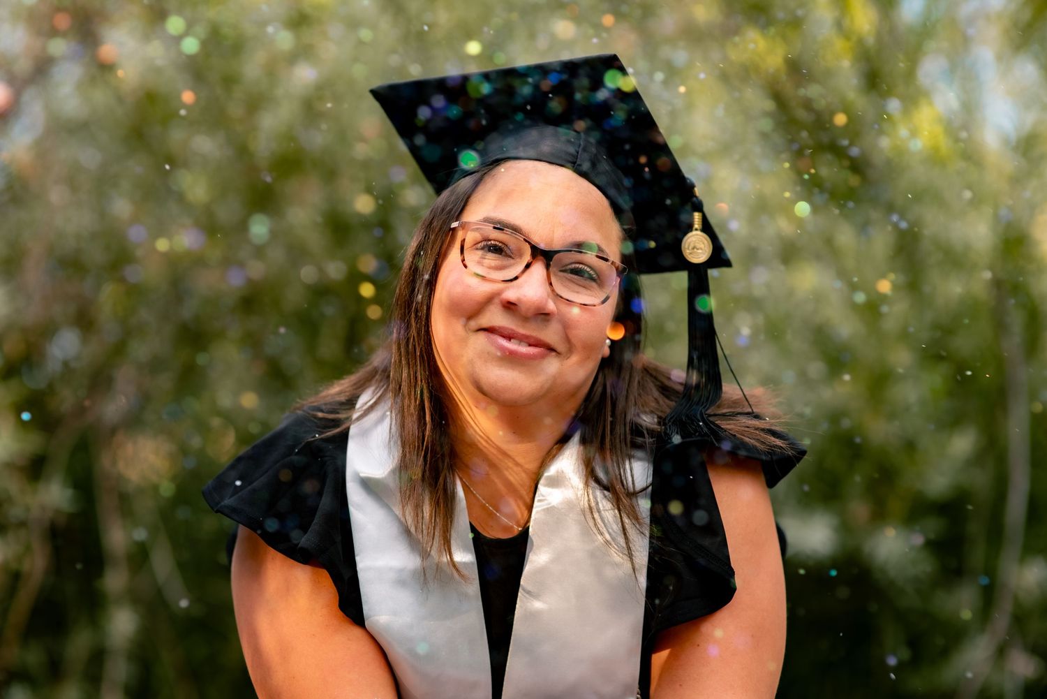 Woman wearing graduation cap with glitter falling through the air creating bokeh in front of the subject with a green background