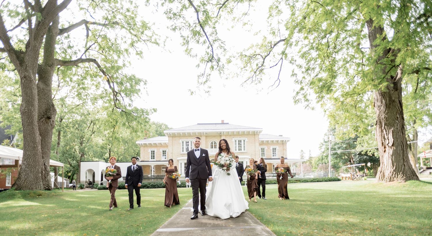 Wedding party gathers under tall trees in a park setting during an outdoor celebration.