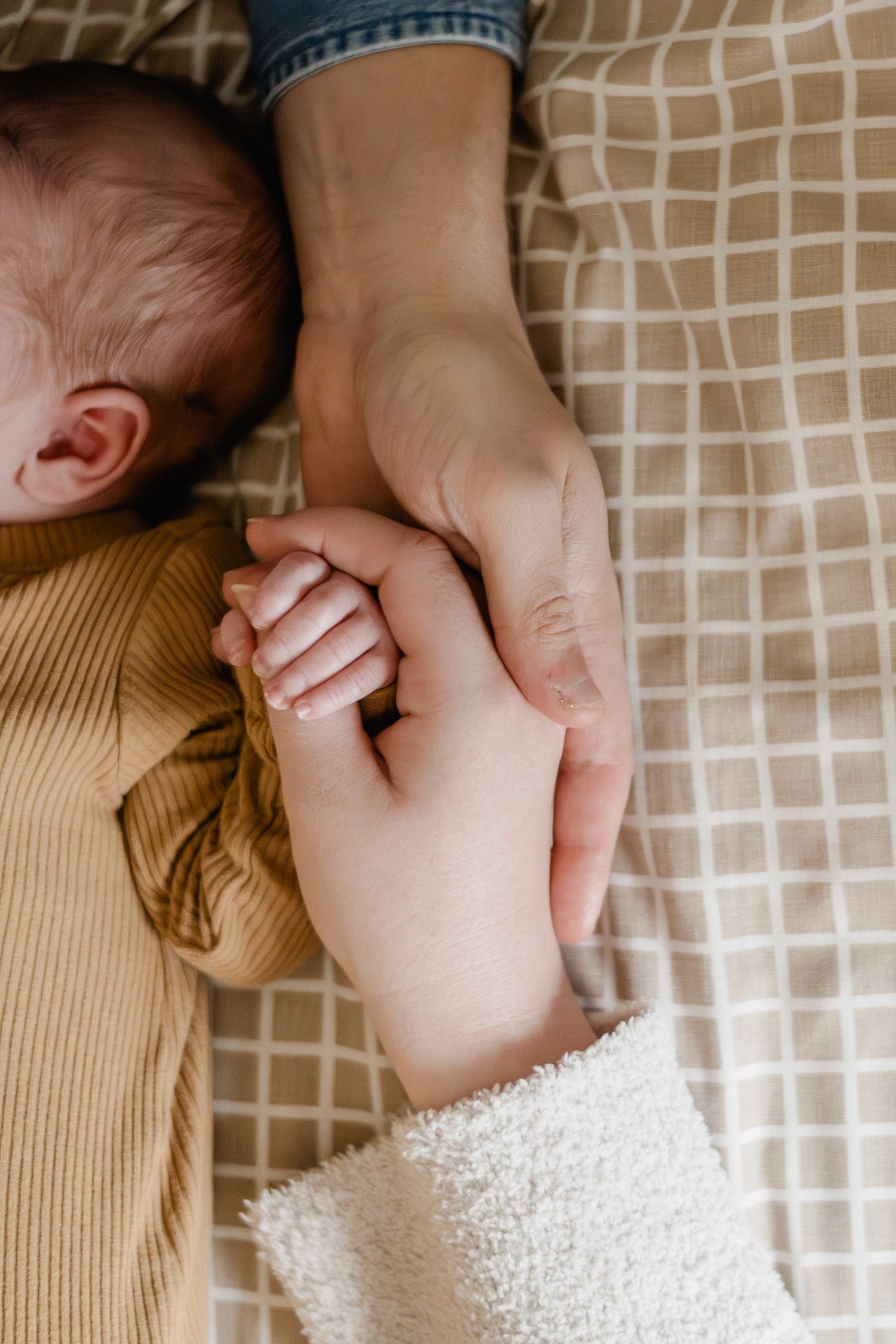 A newborn baby in an orange outfit holds an adult's hand on a beige checkered blanket.