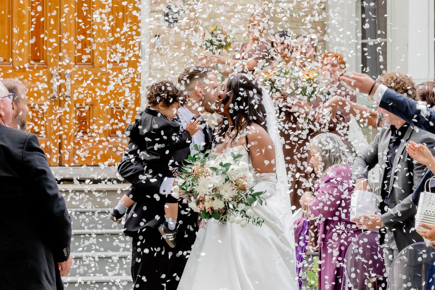 Wedding guests shower the newlyweds with confetti during an outdoor celebration in front of a stone building.
