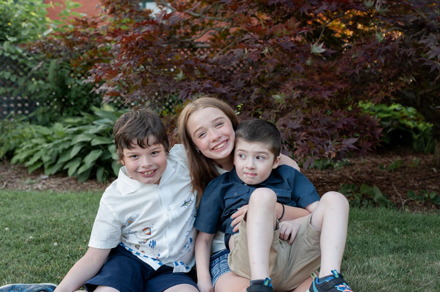 Three people sitting close together and smiling on grass with autumn foliage in the background.