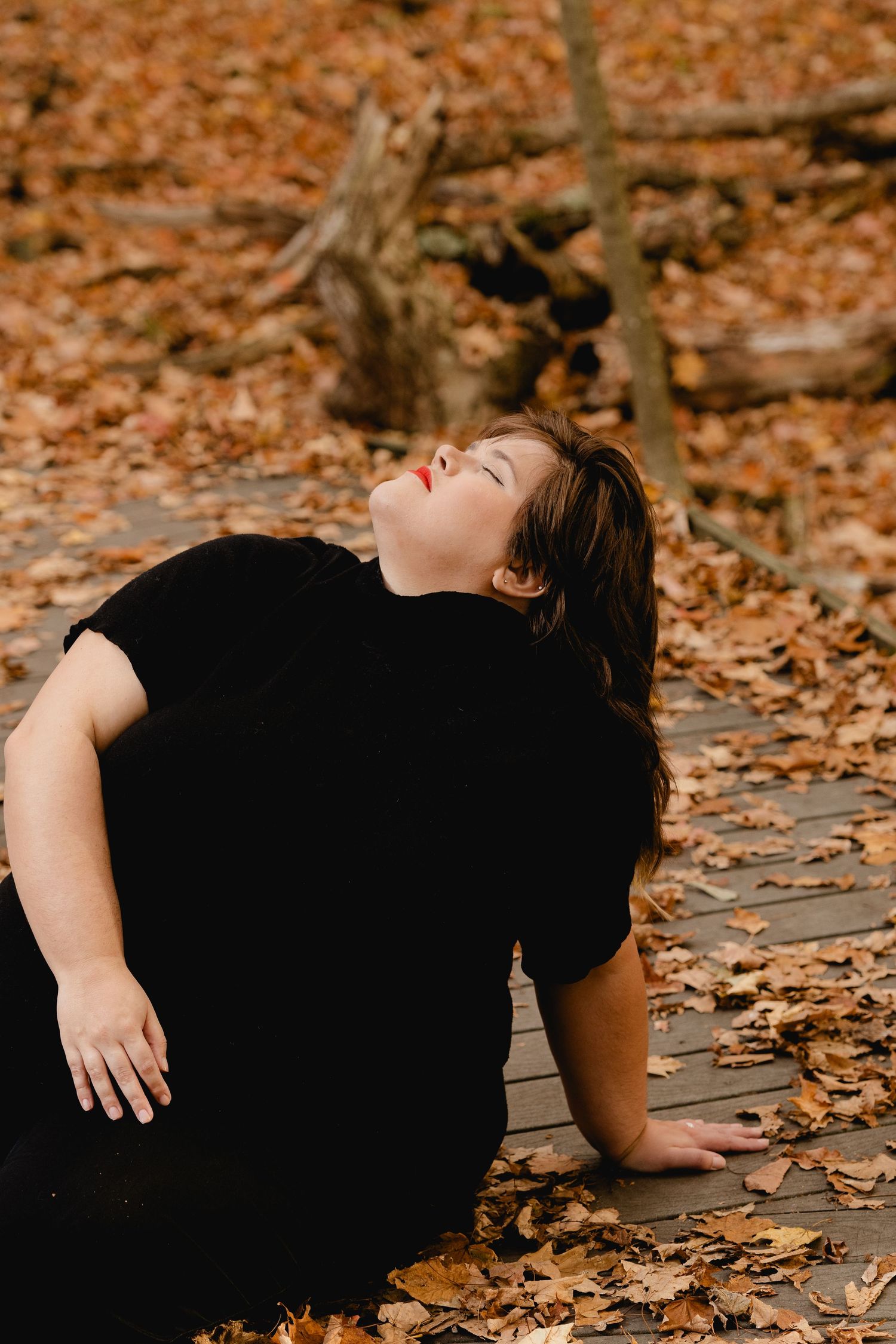 A figure in a black outfit lies dramatically on autumn leaves covering the ground during a photo session.