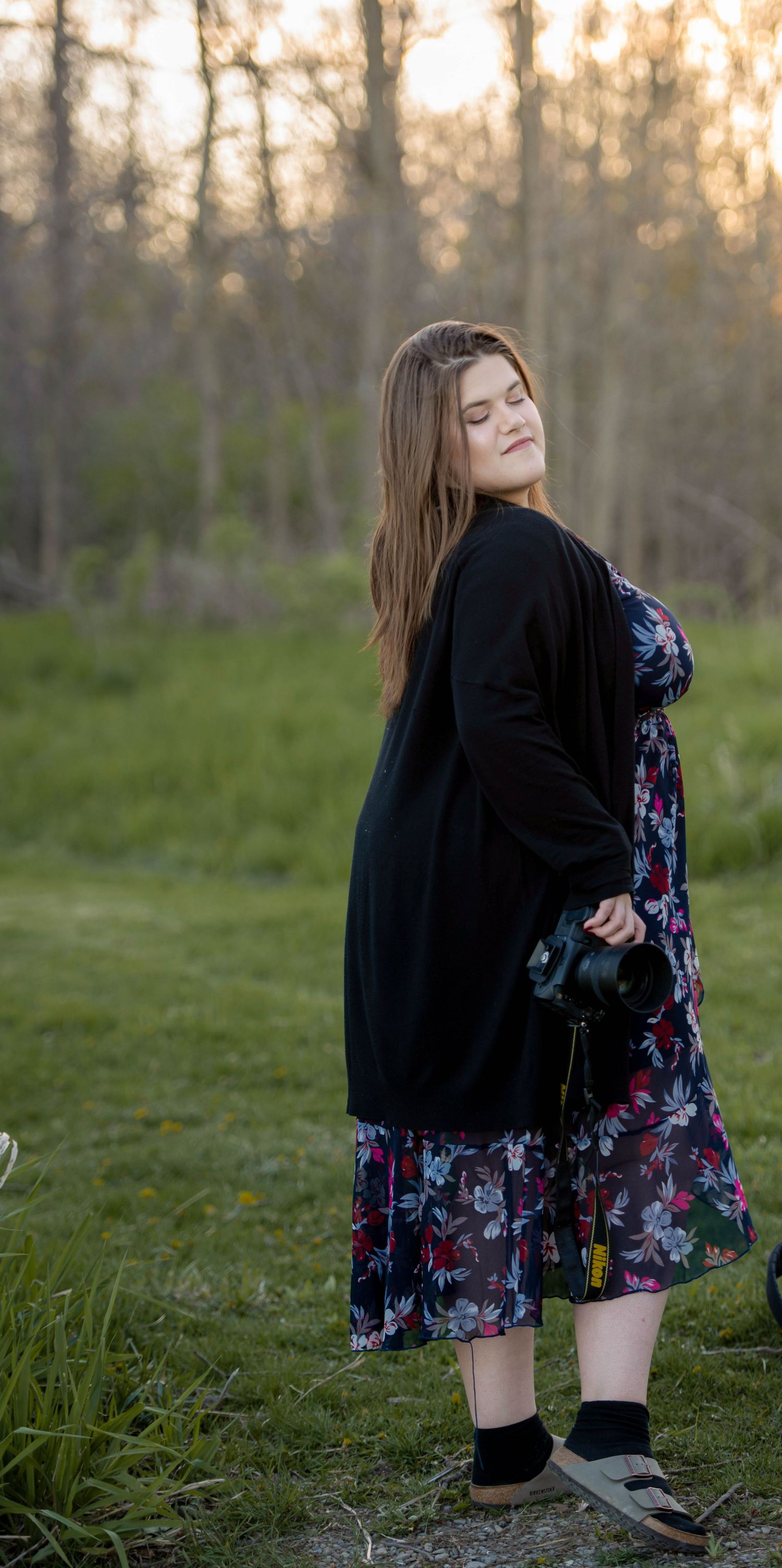 Someone wearing a floral dress and black sweater poses in a natural outdoor setting at dusk.