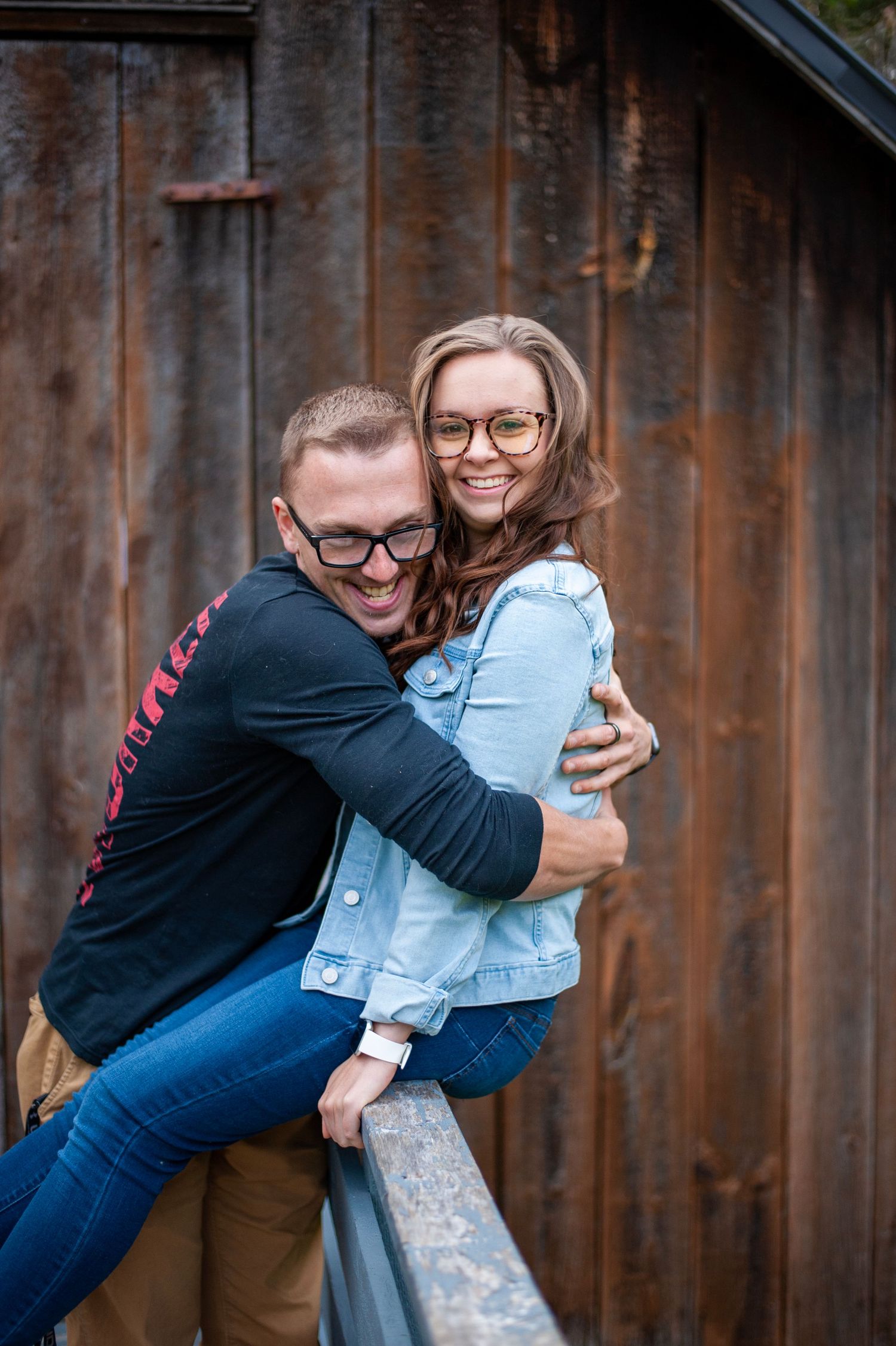 Playful couple pose against rustic wooden barn wall wearing casual denim.
