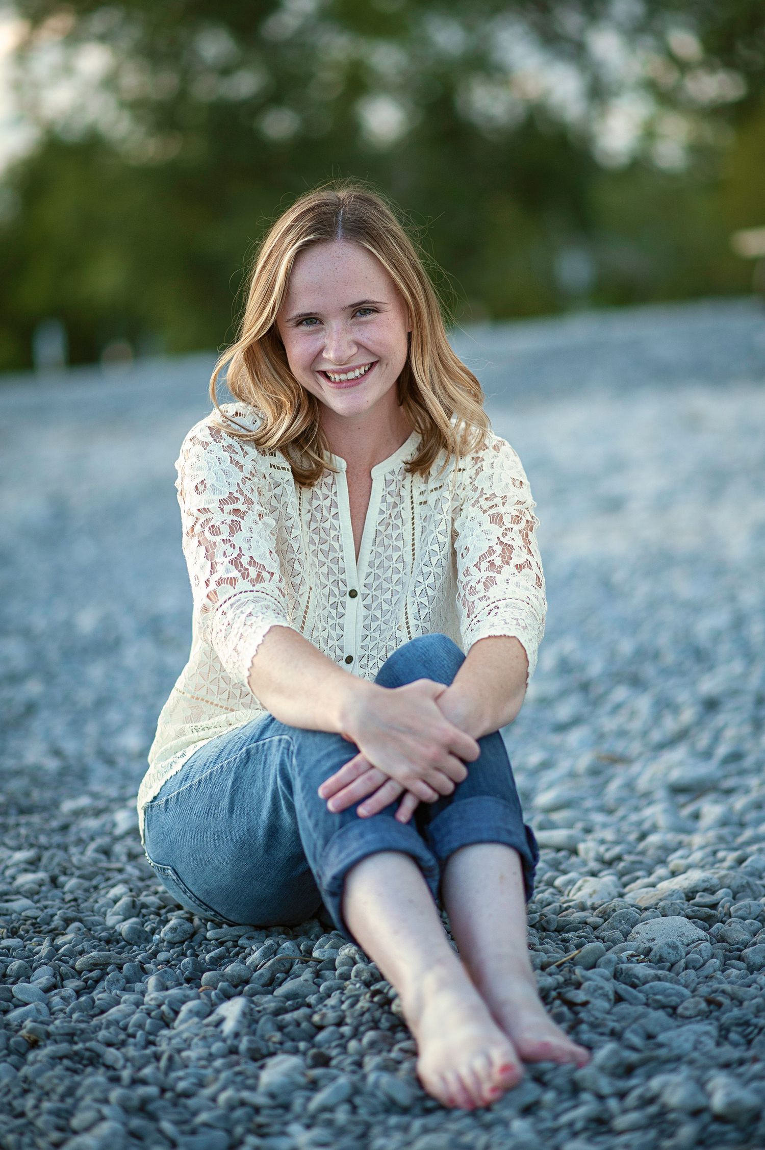 Person in white lace top and jeans sitting on rocky beach.