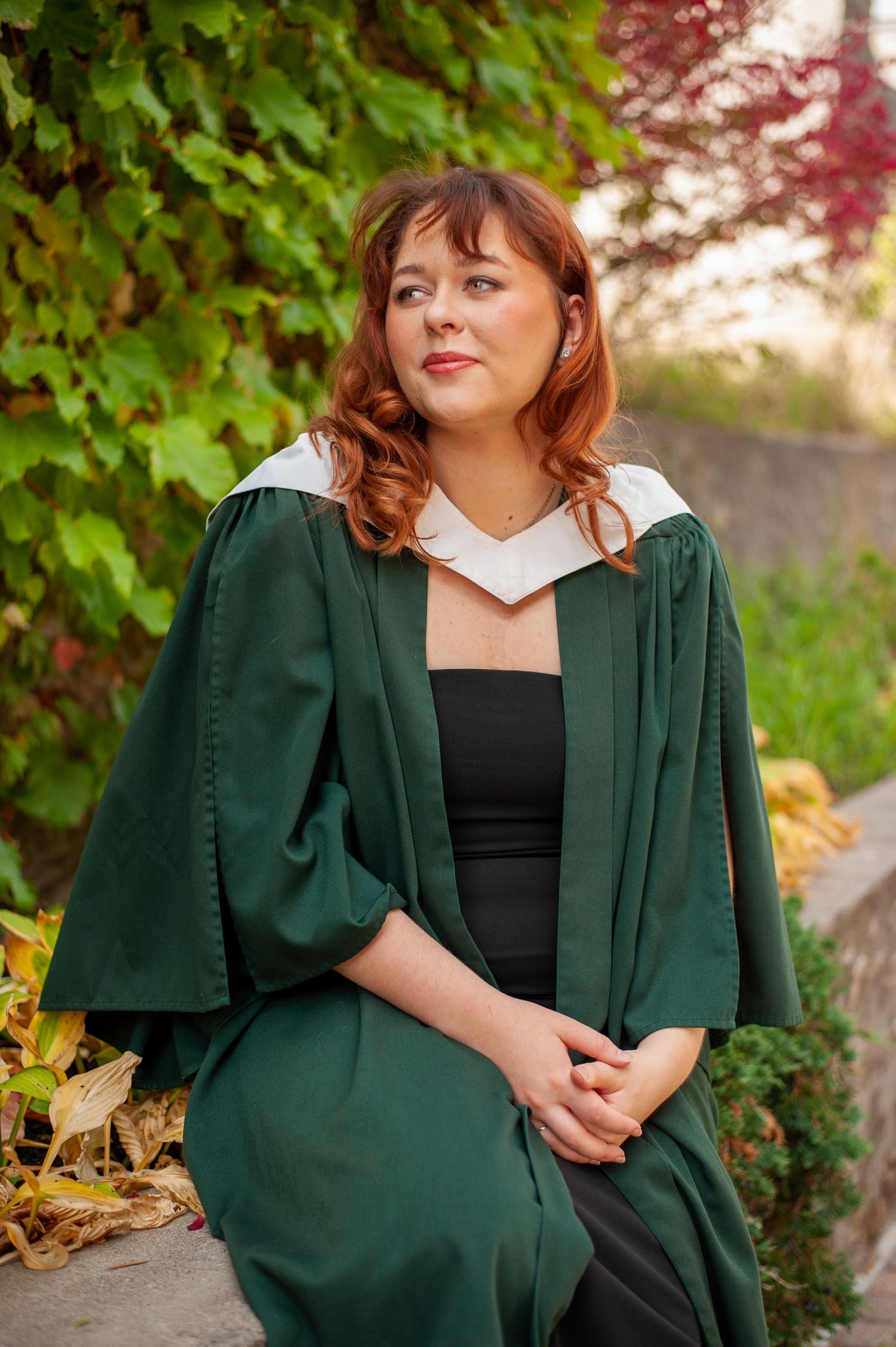 Graduate in emerald green academic regalia poses against autumn foliage.