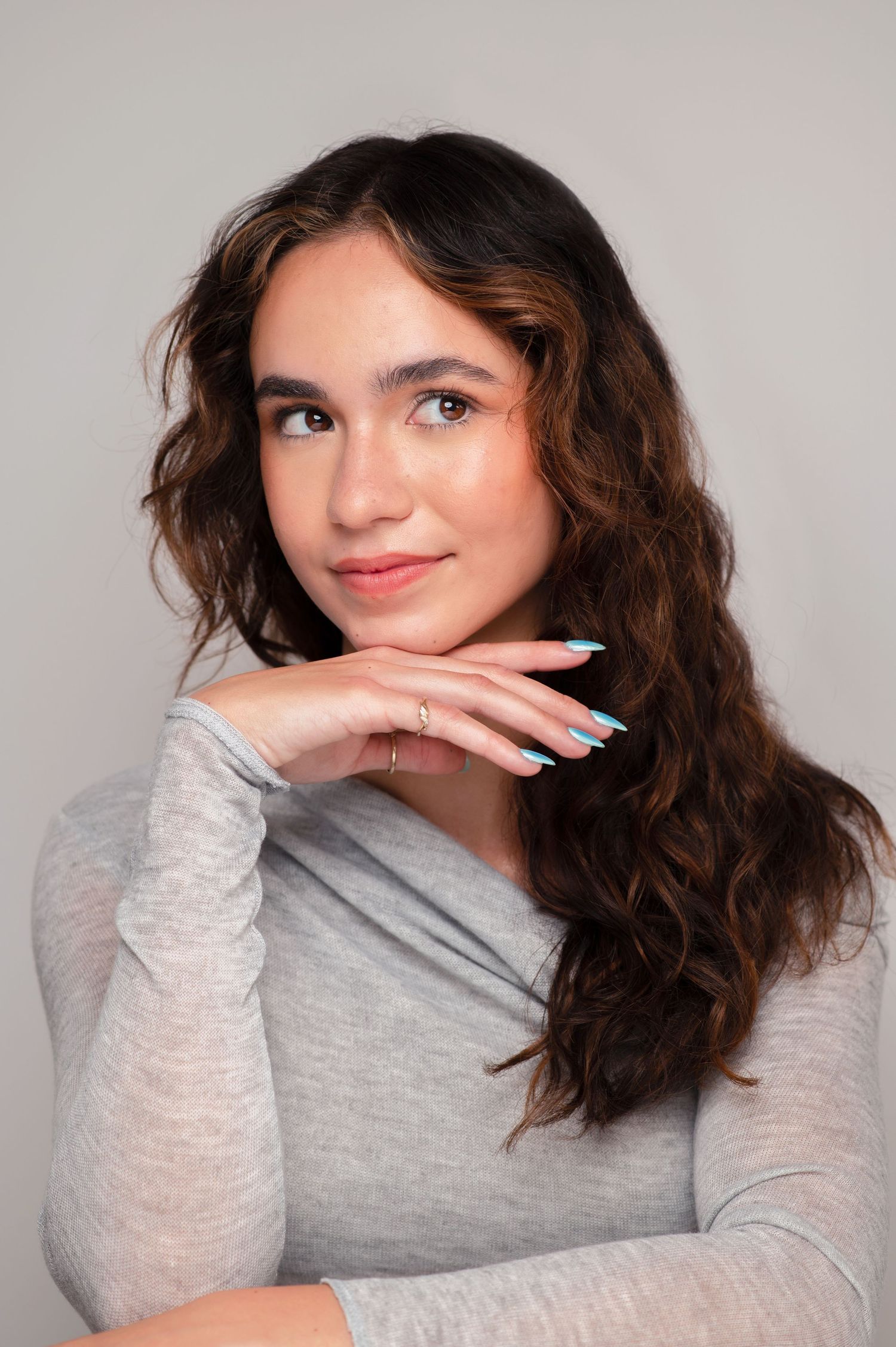 A person with wavy brown hair smiles playfully while wearing a heather grey long sleeve shirt.