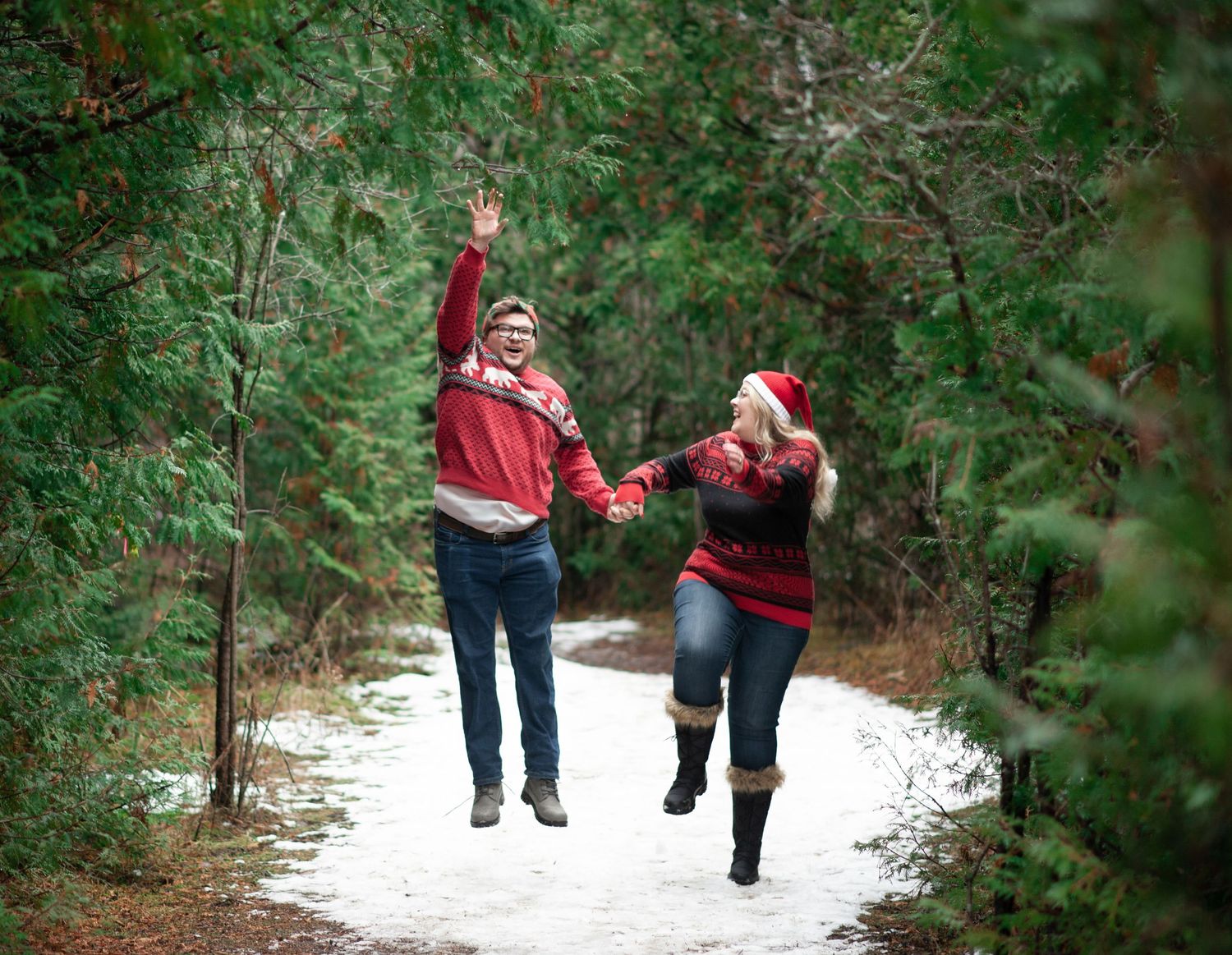A couple in matching red sweaters jumps joyfully on a snowy forest path during winter.