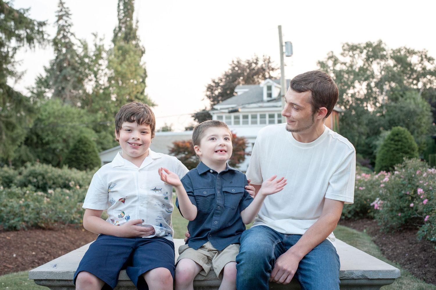Three people share a relaxed moment while sitting together outdoors in a garden setting at dusk.
