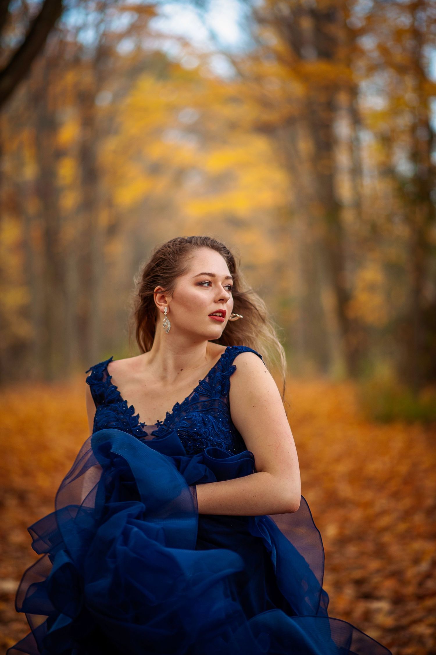 A figure in a flowing navy blue dress poses dramatically among golden autumn leaves in a forest setting.
