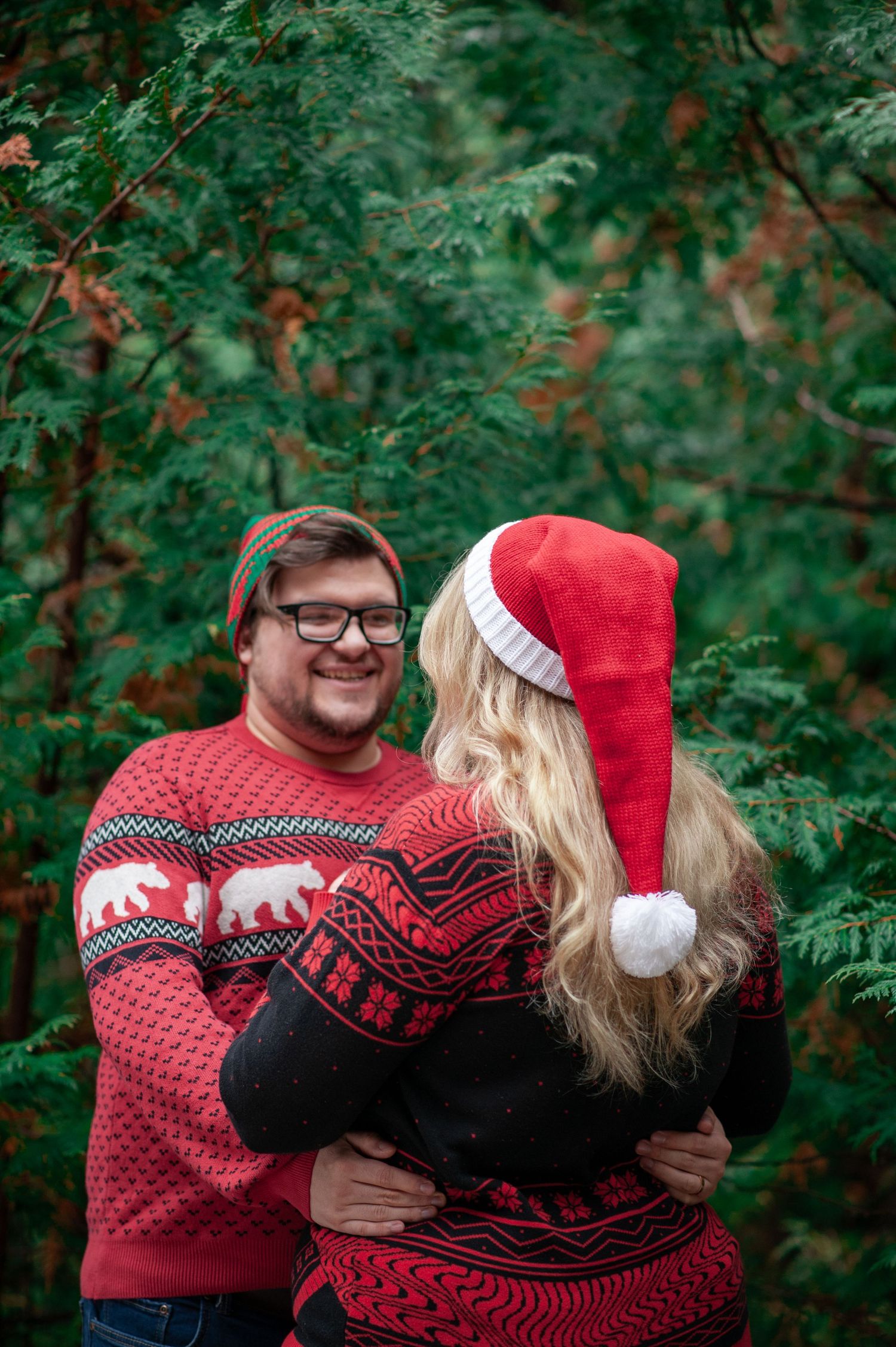 A couple wearing matching holiday sweaters embraces in front of evergreen trees.