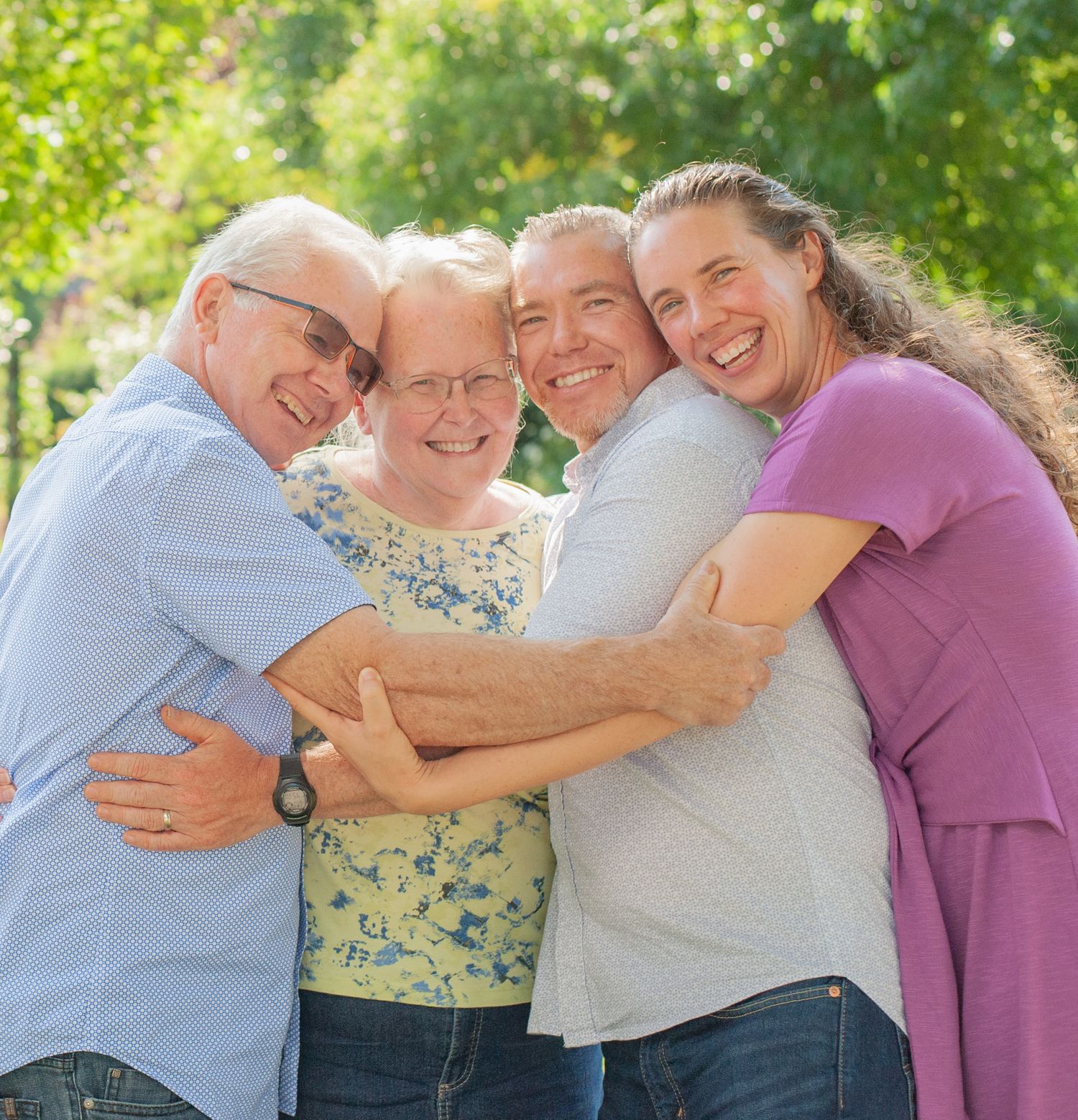 Group of people sharing warm embraces outdoors on a sunny day with green foliage in the background.