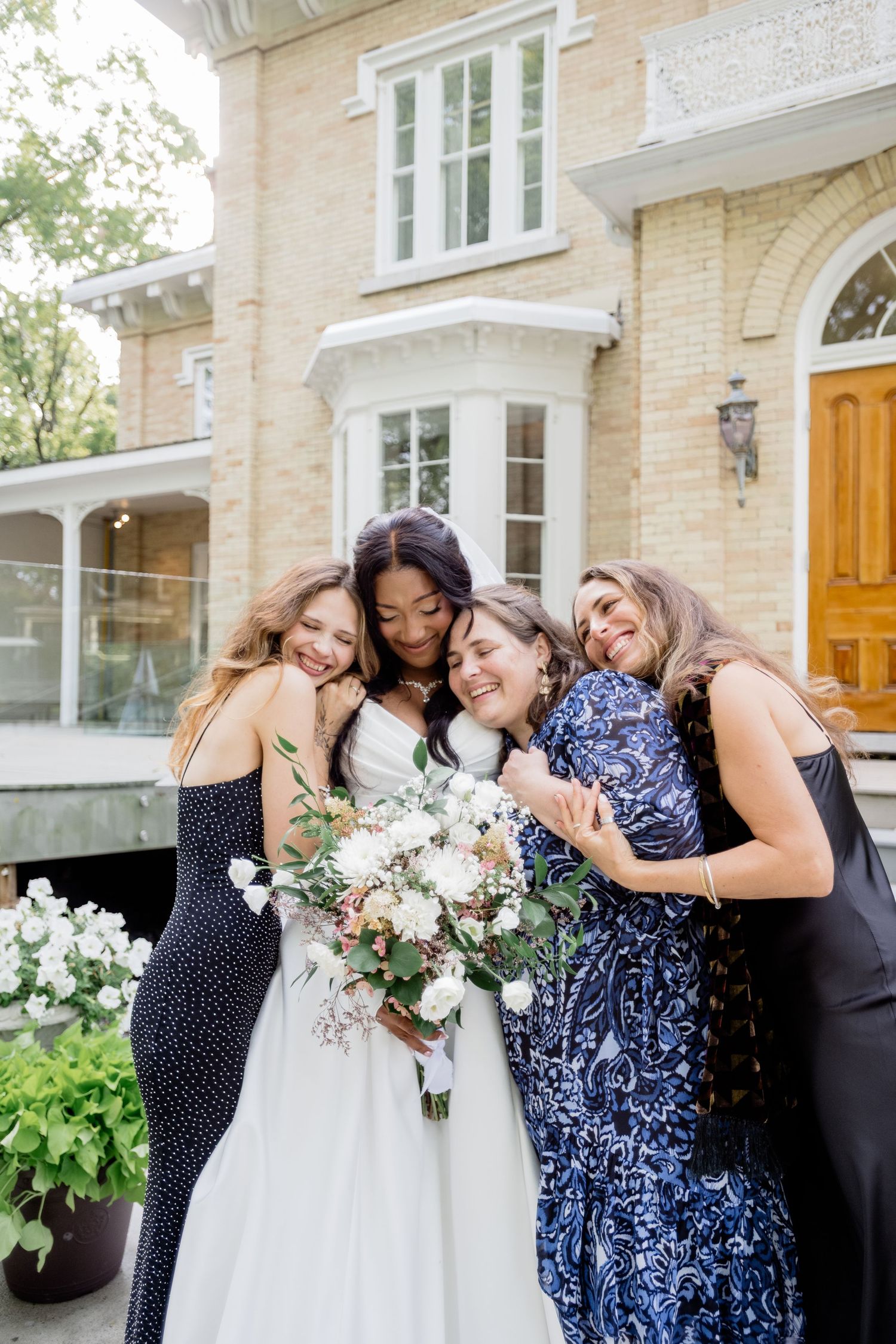 A group of friends in formal dresses share an intimate moment while holding a white floral bouquet outside a brick house.