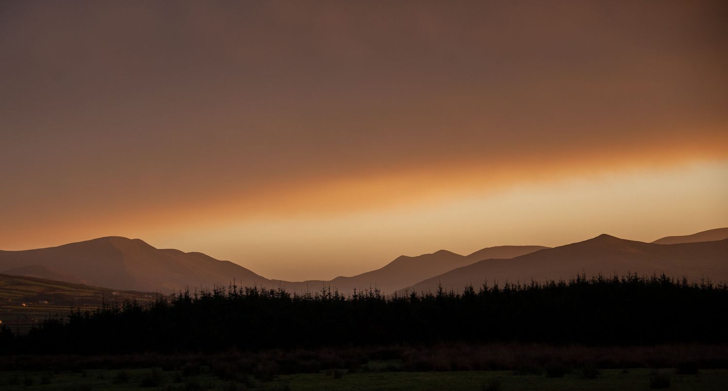 Dramatic sunset sky over mountain silhouettes with forest in foreground.