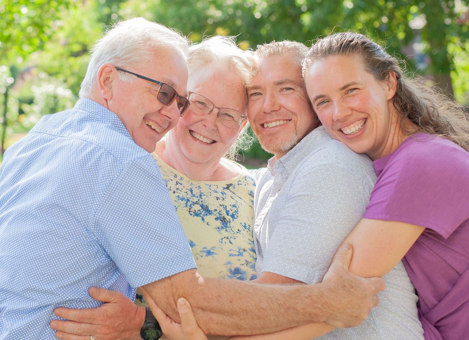 Family members embrace together outdoors on a sunny day in front of green foliage background.