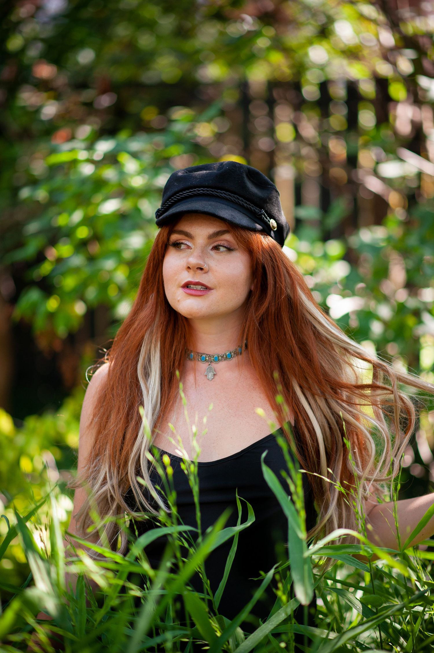 Woman in black dress and newsboy cap poses in lush garden setting.