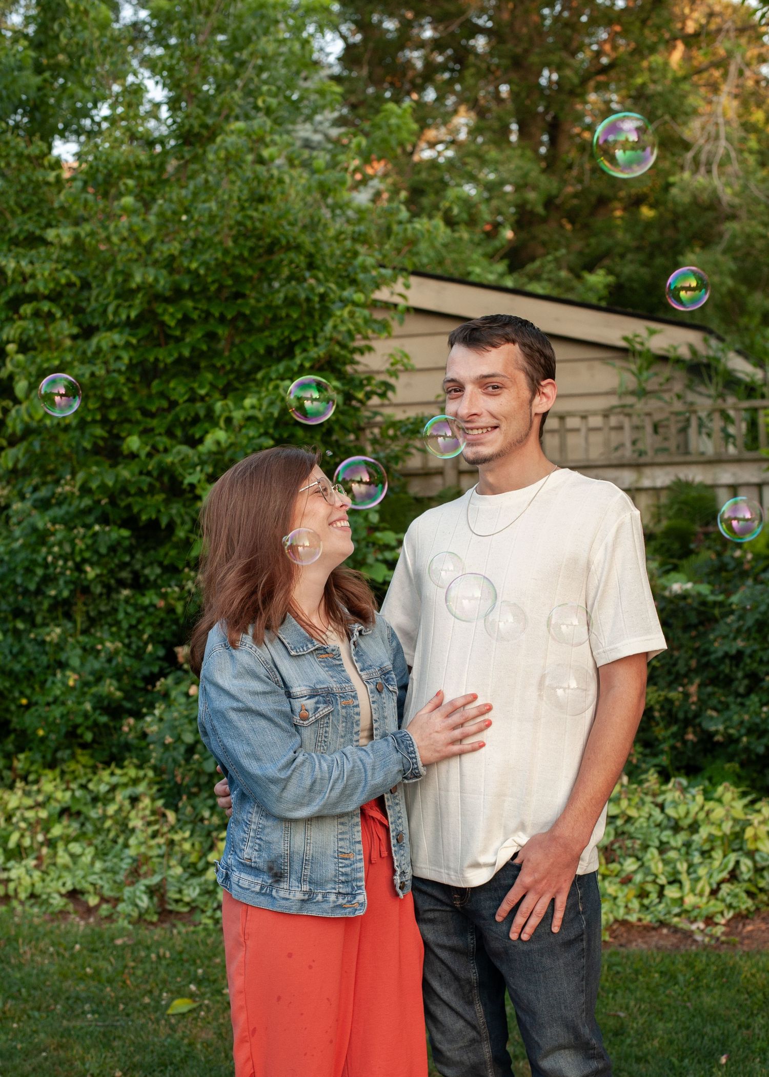 A couple laughs together surrounded by soap bubbles in a garden setting during a casual outdoor photoshoot.