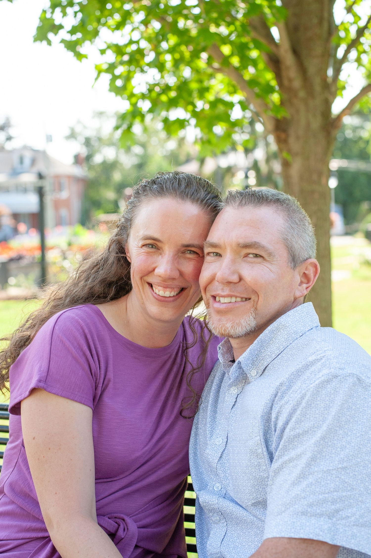A couple sits close together and smiles on a bench in a sunny park setting with trees in the background.
