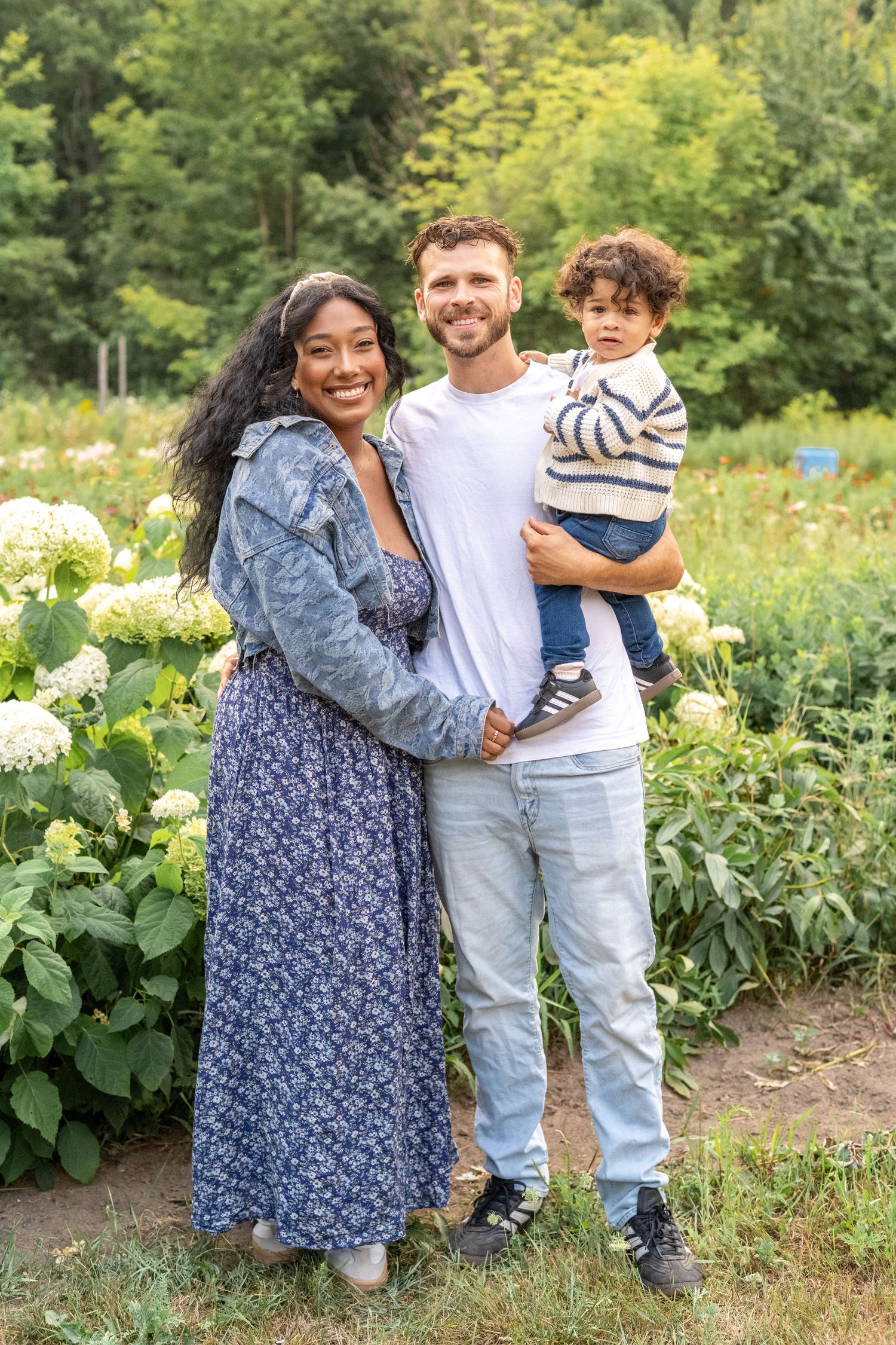 A family poses together in a garden on a sunny day surrounded by lush greenery and white flowers.