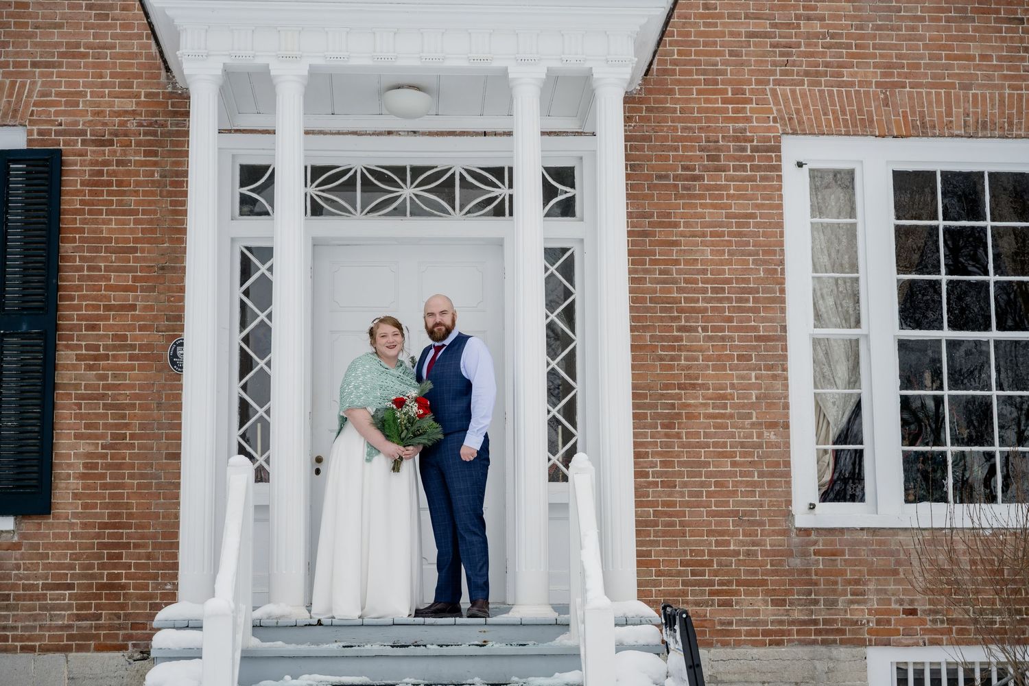 Bride and groom posing on steps of brick building with white columns, bride holding red bouquet in winter wedding photo.