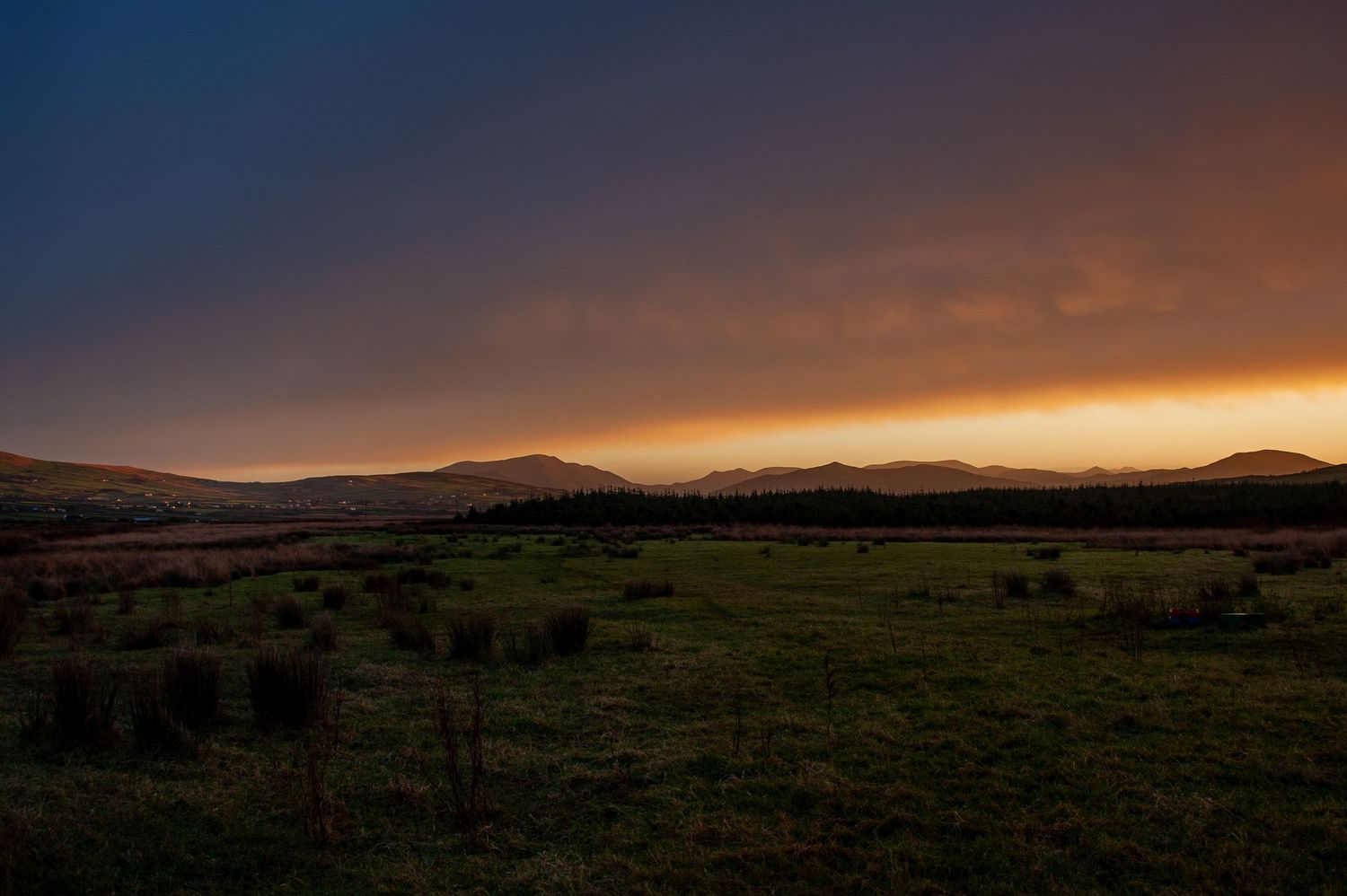 Panoramic sunset view over rolling hills with warm orange sky and dark silhouetted landscape.