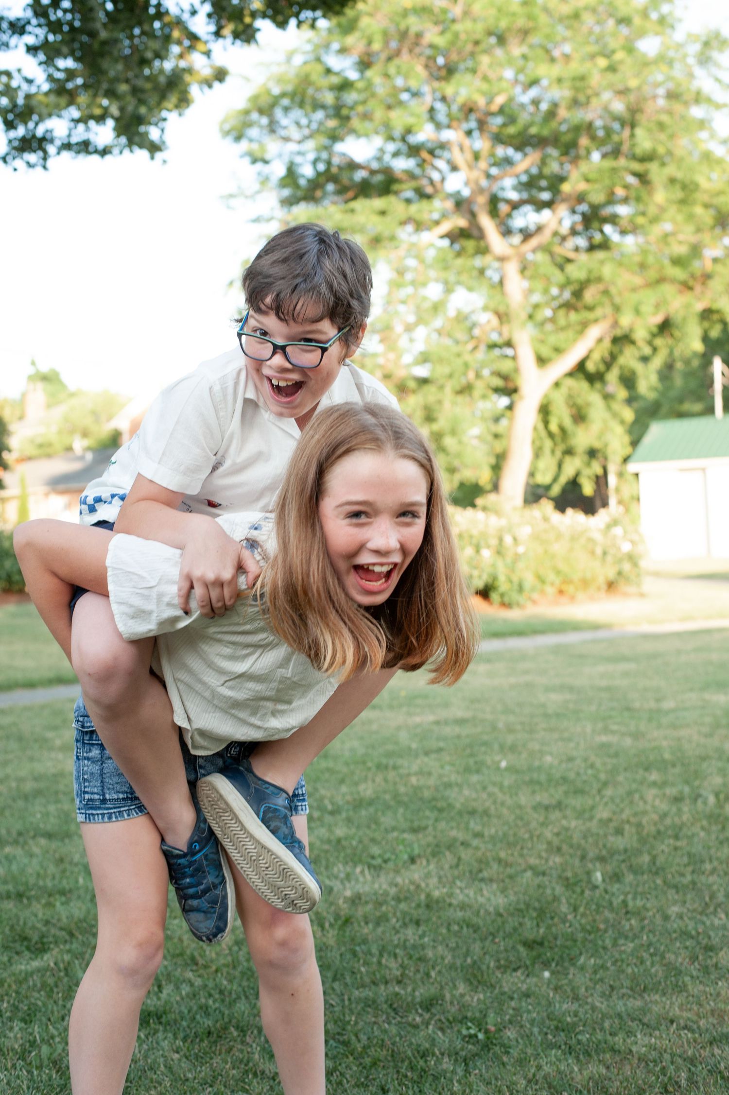 Two kids playing and laughing outdoors on a sunny day in a grassy yard with trees in the background.