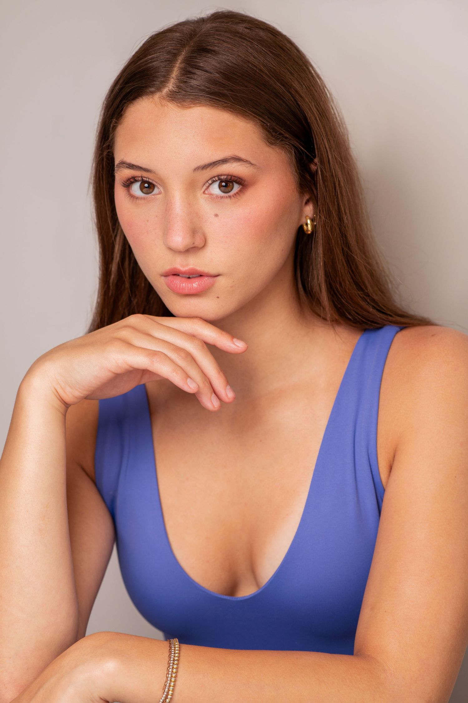 Portrait of a young woman in a blue tank top posing with hand under chin against neutral background.