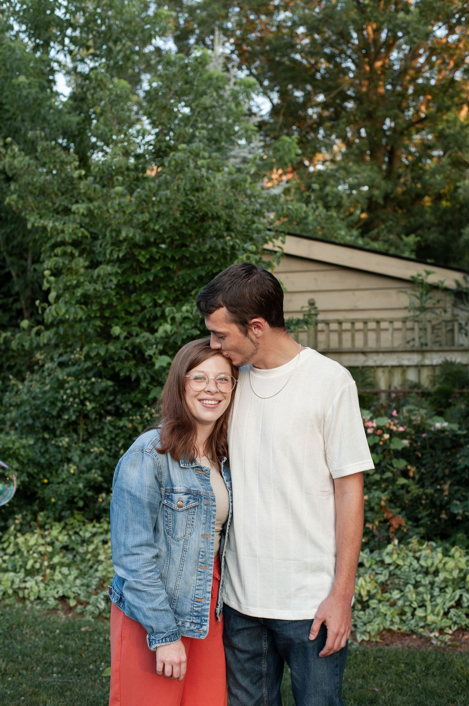 A couple shares a tender moment outdoors, with the person on the right wearing white and the other in denim and coral.